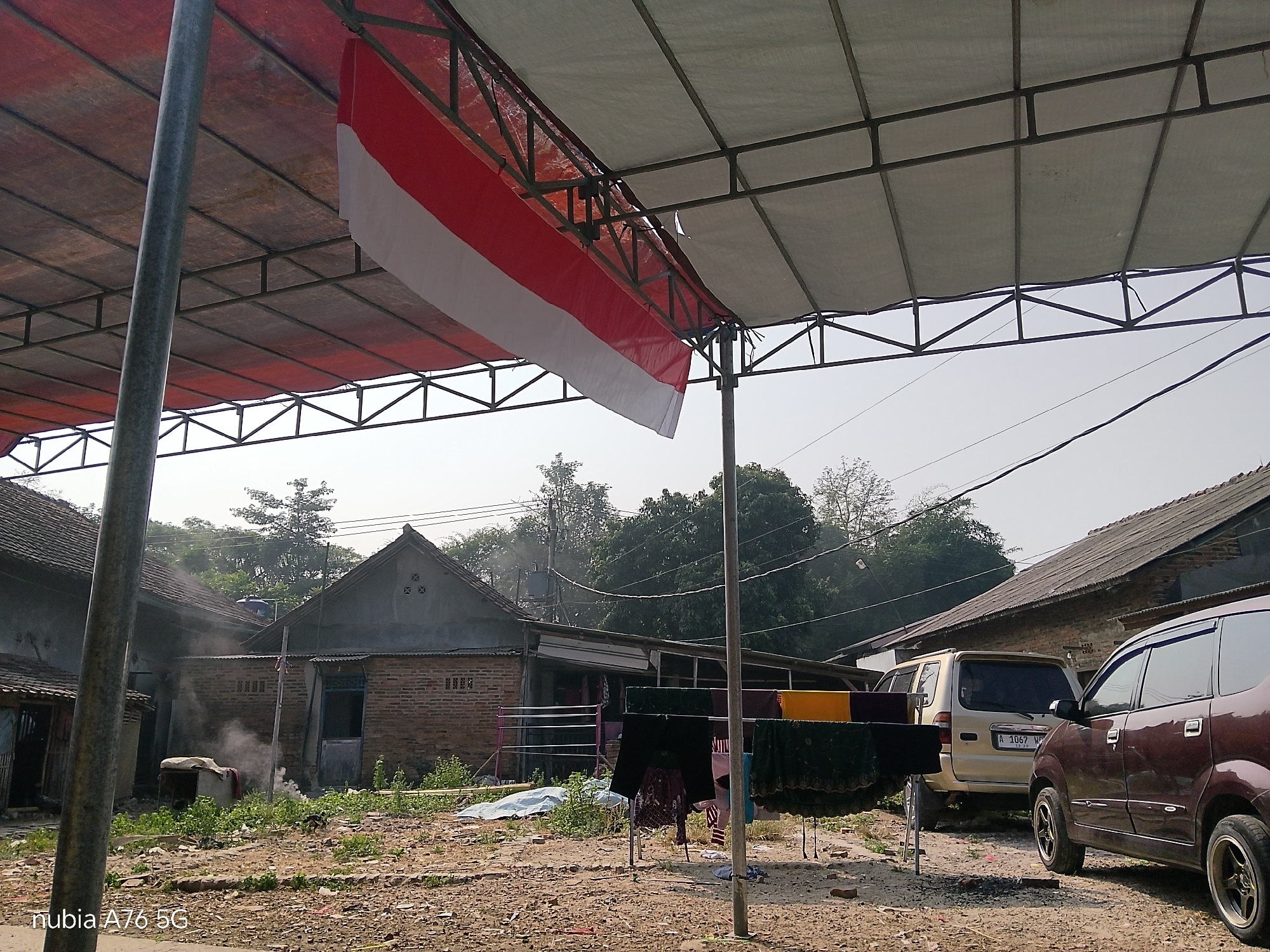 A low-angle outdoor shot captures a scene in Binuang, Indonesia, under a clear, overcast sky. A red and white Indonesian flag hangs diagonally from a metal frame supporting a large, taut fabric canopy. The canopy itself is composed of two sections: the left side is a reddish-brown fabric, while the right side is a lighter, off-white material. Both sections are supported by a dark grey metal framework.
To the left, a tall, grey metal pole stands vertically, partially obscuring the view. Beyond the pole, a rustic building with a tiled roof and brick walls is visible. Smoke or steam emanates from a small, enclosed area in front of this building, surrounding a rectangular object covered by a cloth. Green vegetation grows in patches around the building and across the ground, which is a mix of dirt, gravel, and scattered debris.
In the mid-ground, a clothesline supports several pieces of laundry, including dark-colored garments and yellow and green fabrics. A pink drying rack is also visible behind the laundry. Further to the right, two vehicles are parked. The one on the left is a light-colored SUV, and the one on the right is a darker maroon minivan. The minivan's license plate is partially visible. Behind the vehicles and laundry, more buildings with corrugated metal or tiled roofs are present, along with numerous trees. The ground is uneven and appears to be a dirt or gravel path. A watermark in the bottom left corner reads "nubia A76 5G".