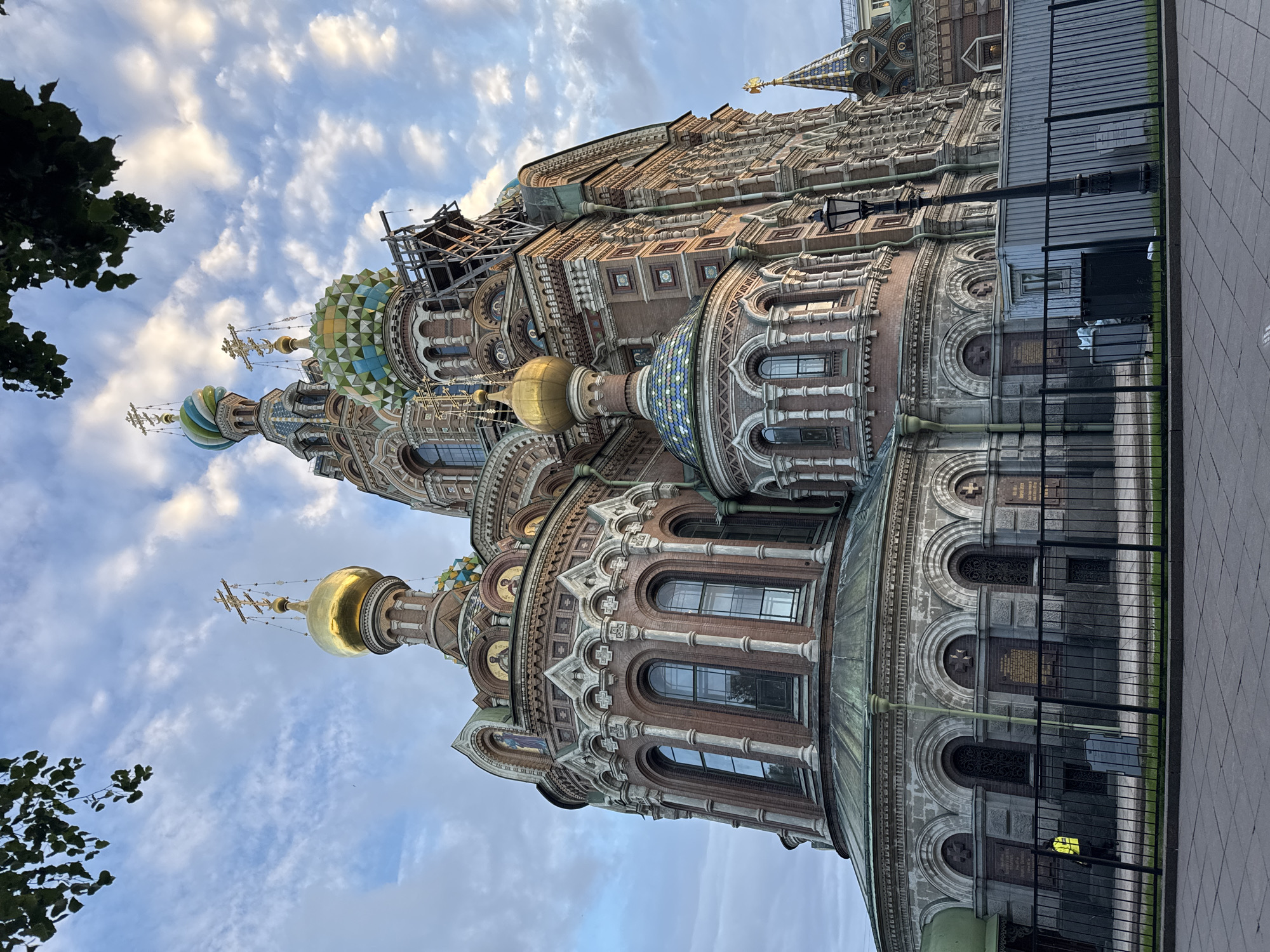 The image shows the Church of the Savior on Spilled Blood, a prominent landmark in Saint Petersburg, Russia. The church is viewed from a low angle, looking up at its ornate architecture and colorful domes against a cloudy sky. The building features red brick walls, white decorative elements, and multiple domes, some of which are gilded and others are multicolored with intricate mosaic patterns. Scaffolding is visible on one of the domes, suggesting ongoing restoration work.
In the foreground, a black metal fence encloses the church grounds. Behind the fence, there is a paved walkway and some greenery. To the left of the church, a person wearing a bright yellow vest is visible behind the fence. A metal shed and some trash bins are also present behind the fence. A lamppost stands to the right of the shed, in front of the church wall. The overall scene captures the grandeur of the church under a dynamic sky, with some elements of urban infrastructure and human presence.