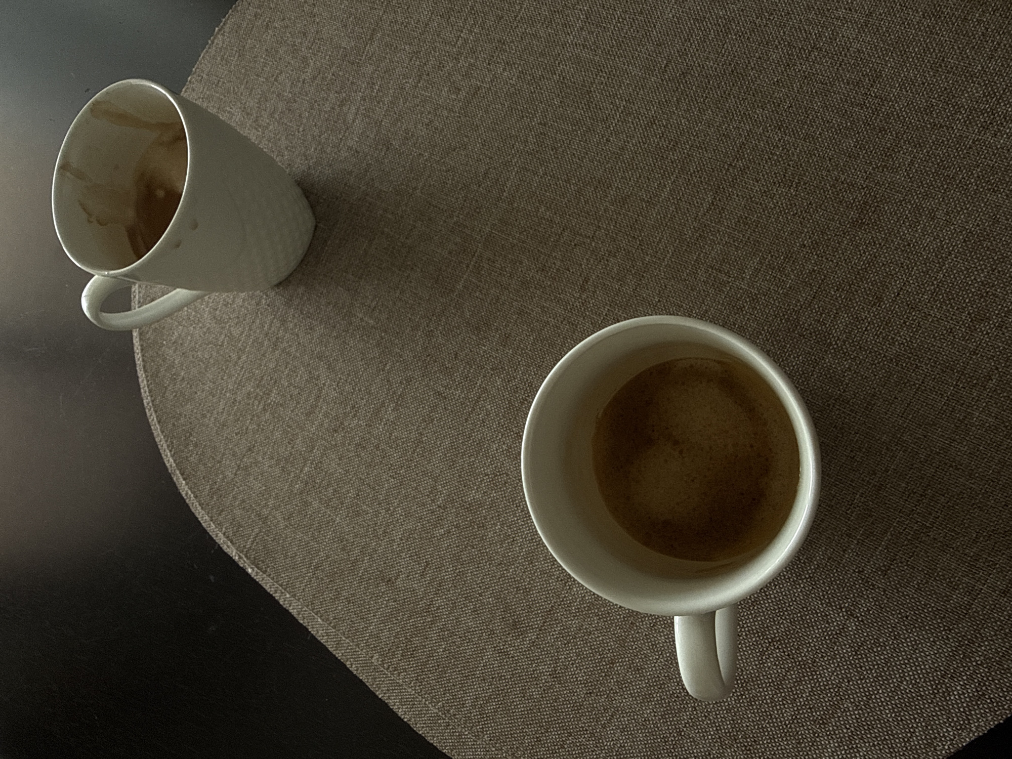 The image shows two white ceramic mugs of coffee on a brown textured placemat. One mug is in the foreground on the left, and the other is in the background on the right. The mugs are tilted, and the coffee inside is dark brown with some foam. The background is a dark surface, likely a table. The lighting suggests an indoor setting. The image does not contain any information or context that would allow for determining a location in the city of Nesebar, Bulgaria.