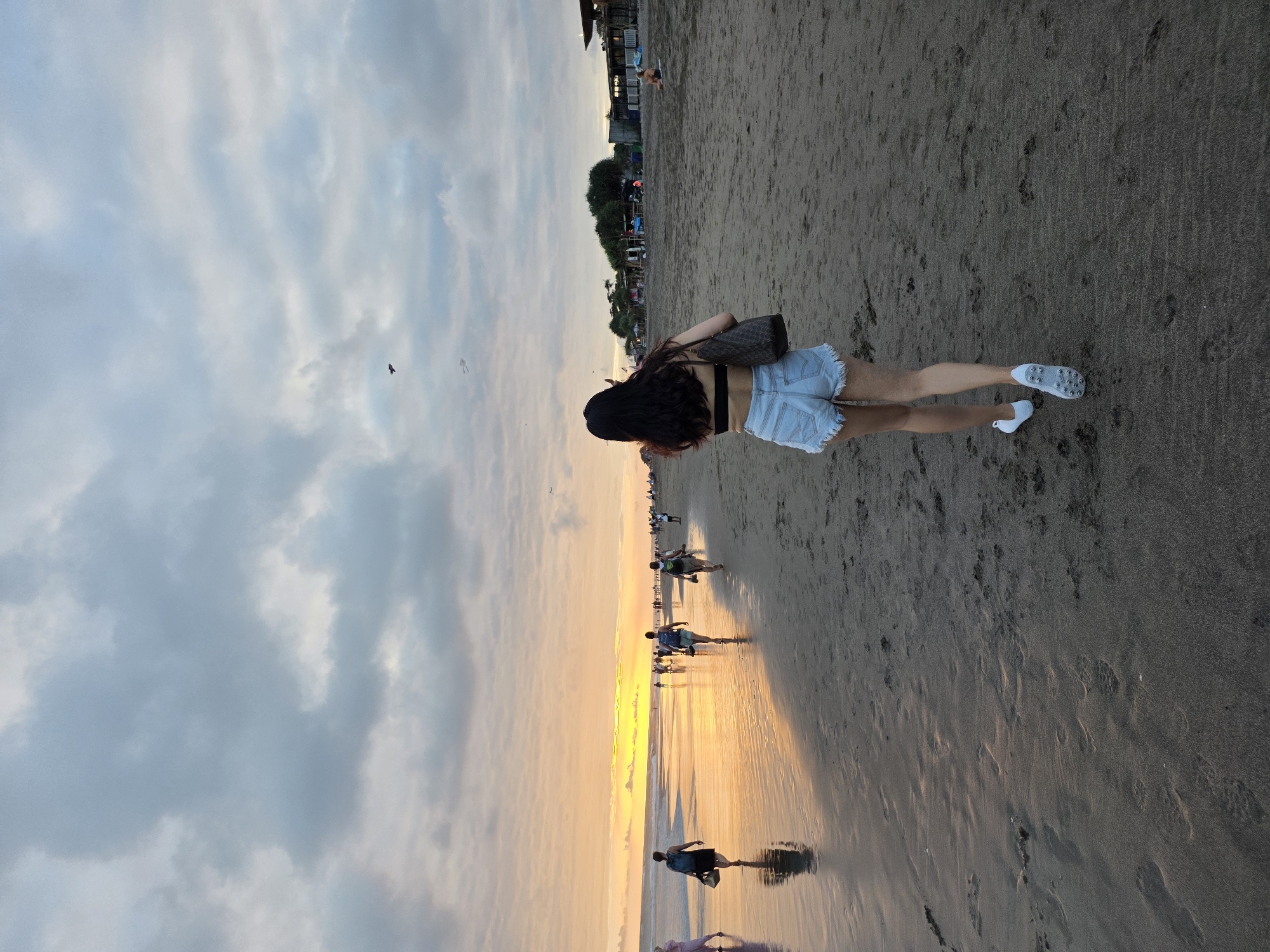 The image depicts a beach scene in North Kuta, Indonesia, during twilight. The sky is filled with dramatic clouds, with the setting sun casting a warm orange and yellow glow on the horizon. A woman, seen from behind, is walking away from the camera on the sandy beach. She is wearing denim shorts, a black bikini top, and white sneakers. She carries a dark brown tote bag over her shoulder. In the background, other people are walking along the shoreline, some closer to the water's edge, and a few buildings are visible further inland. The wet sand reflects the sky's colors. Two kites are visible flying in the distance.