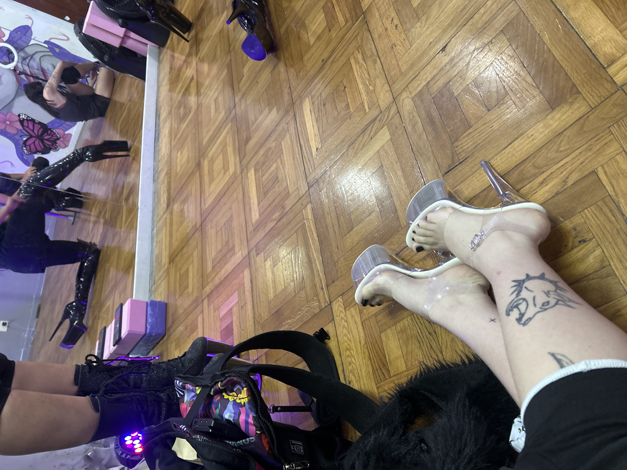 This elevated shot captures an indoor scene with a wooden parquet floor, likely a studio or practice space in Buenos Aires, Argentina.
In the foreground right, a person's bare legs and feet are visible, wearing clear high-heeled sandals with black toenail polish. Tattoos adorn the right leg: a small 'x' near the ankle and a distinctive stylized animal skull (resembling a horse or donkey) with sharp teeth and a spiky mane on the calf.
To the foreground left, another person's legs are seen in black shorts and black, possibly glittery, lace-up boots. A black backpack with a vibrant, colorful abstract pattern on its front pocket rests nearby, next to a black furry object. A device emitting a purple light is partially visible beside the backpack.
A large mirror or reflective surface along the left side of the frame provides a wider view of the room and its occupants. Reflected elements include a person in black shorts and top, a shiny black long glove or sleeve on another person's arm, a pair of very tall, shiny black platform boots, stacked pink and purple blocks (possibly yoga props), a black fan, and a ring light.
The scene suggests a moment of rest or preparation, likely associated with performance art such as pole dancing or drag, given the specialized footwear and studio equipment. The environment appears well-lit. No discernible text is visible in the image.