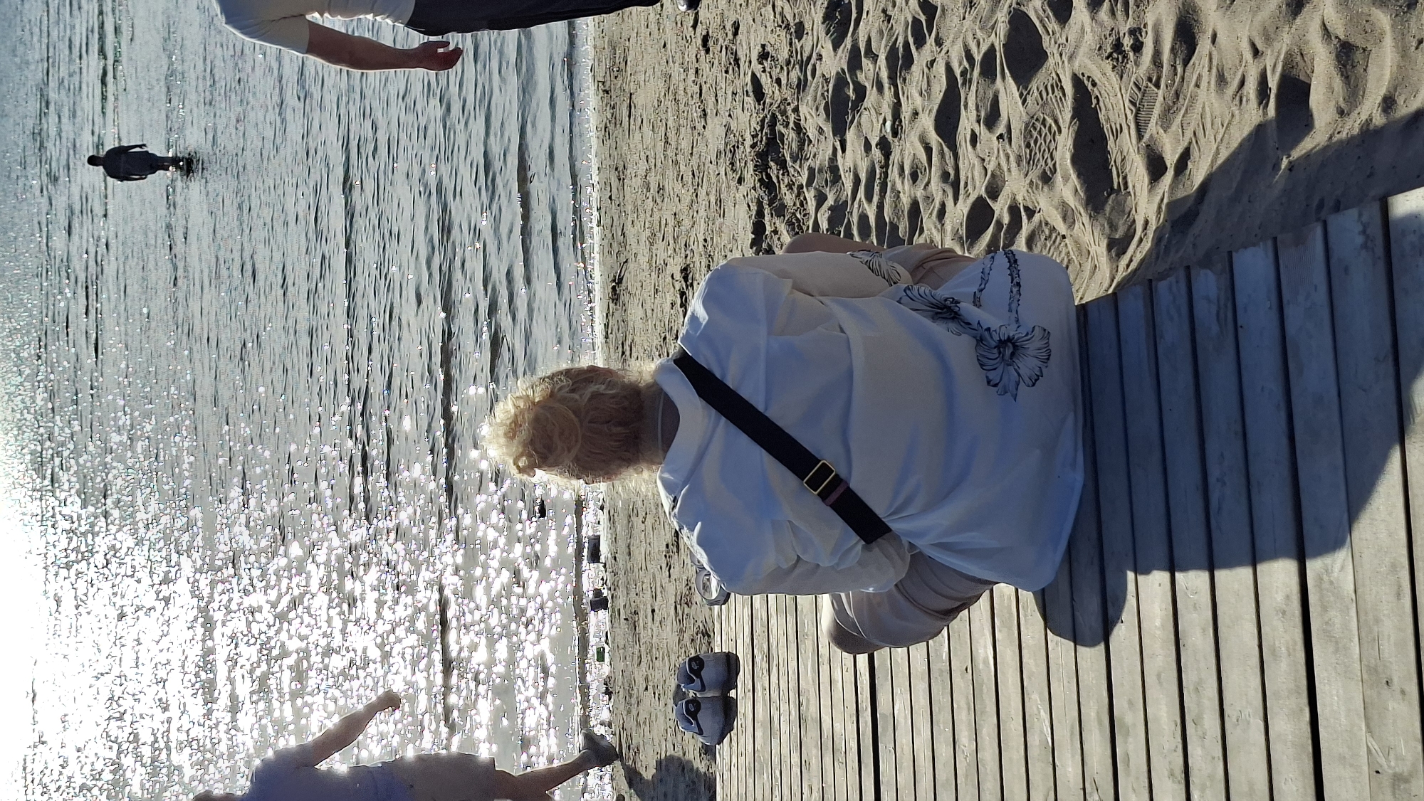 A sunny day scene at the Tallinn beach shows several people. In the foreground, a woman with blonde hair pulled back in a bun sits facing away from the camera on a wooden boardwalk. She is wearing a white button-up shirt and light-colored pants, and a dark strap with a buckle is slung across her chest. She has a white purse with a floral design on her lap. To her left on the boardwalk are a pair of white sneakers. Further out in the shimmering water, a man is wading. In the background, another person stands partially in the water on the left, and a third person's torso is visible on the right, out of focus. The beach sand is visible in the middle ground.