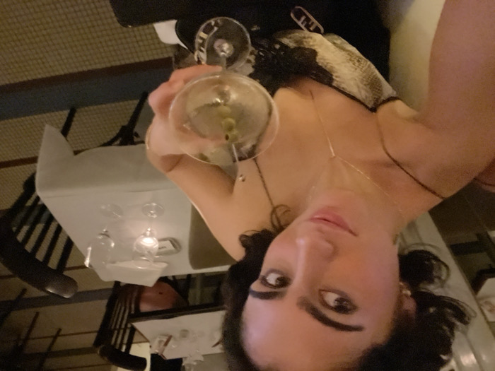 A woman poses with a martini glass filled with olives, wearing a black dress with gold jewelry. She is sitting at a restaurant table with a white tablecloth, multiple wine glasses, and a candle. The restaurant's decor includes dark chairs and a patterned floor. The image is taken from a slightly elevated angle, focusing on the woman's face and the cocktail.
