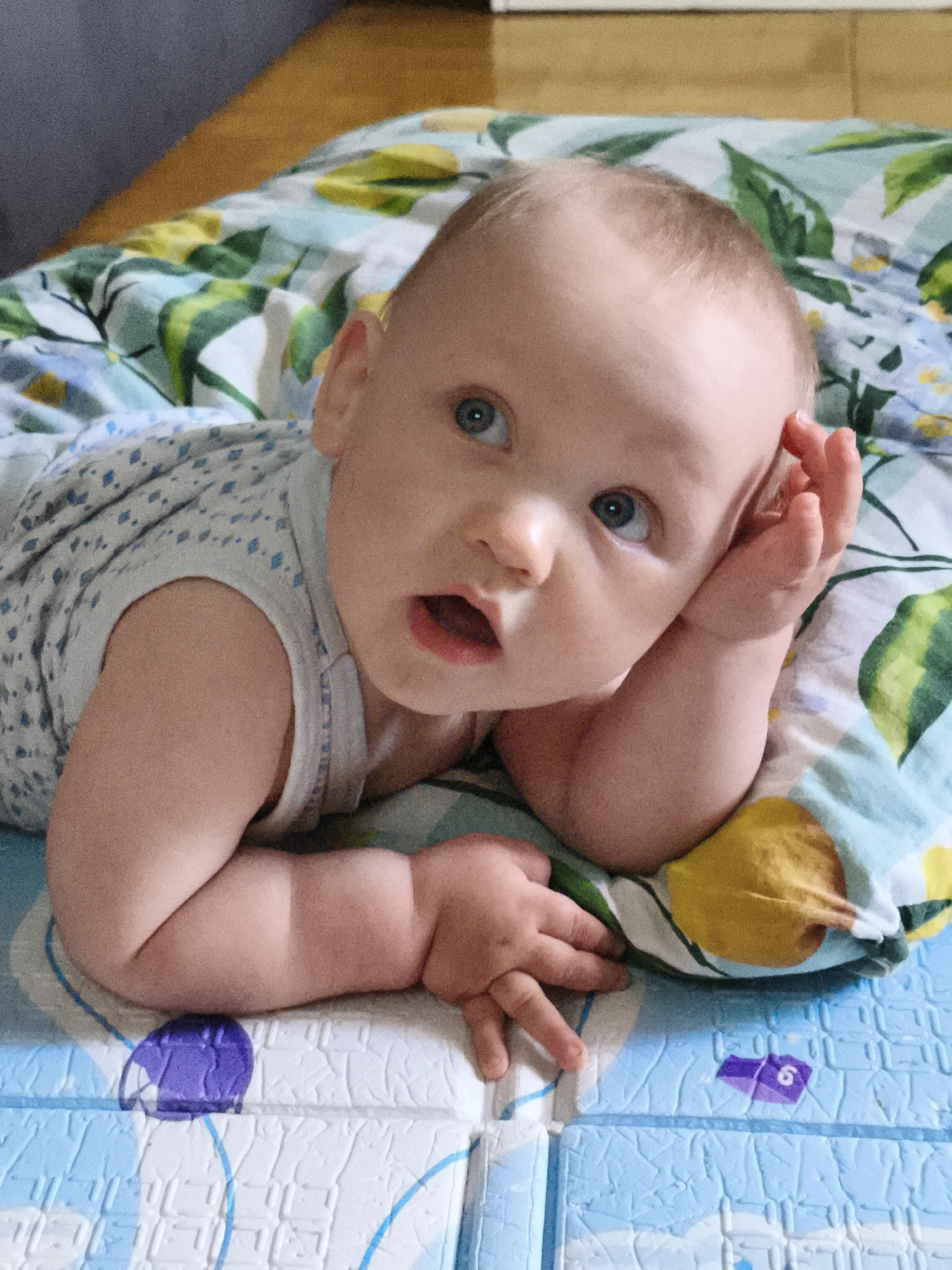 A baby is lying on its stomach on a blanket with a floral pattern. The baby has light hair and blue eyes and is looking upwards and to the right. Its left arm is bent, with its hand resting on its forehead. Its right arm is extended in front of it, with its hand resting on the blanket. The baby is wearing a white and blue patterned onesie. The blanket is on a blue and white mat with a textured pattern. The background is blurred and appears to be a wall and furniture. The image was taken in Chaikovsky, Russia.