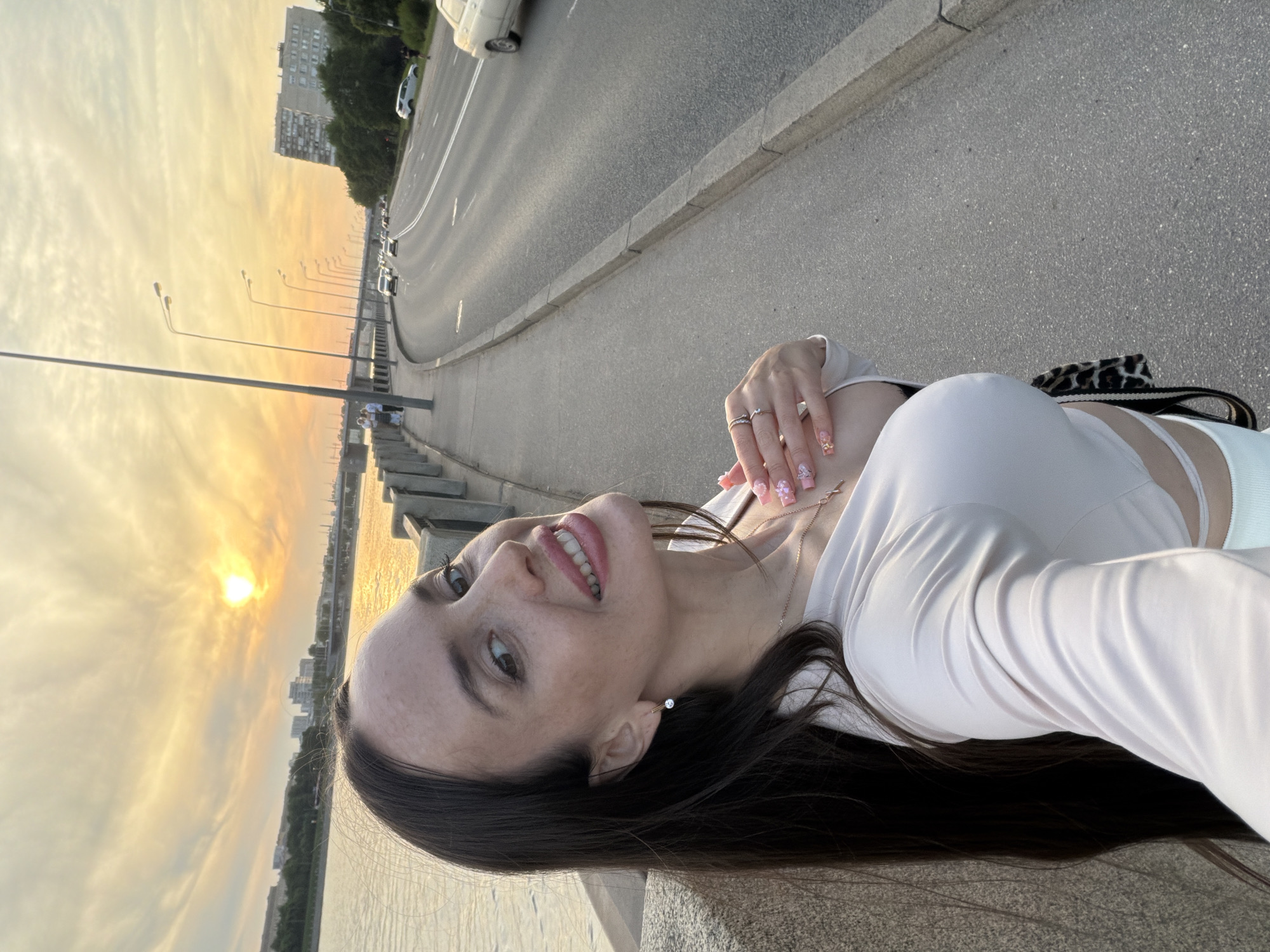 A selfie-style portrait captures a smiling woman on a city embankment during sunset in a city in Unknown, Russia. The woman, with long, dark brown hair and visible freckles, smiles towards the camera with pink-painted lips. She wears a light-colored, long-sleeved crop top, a small stud earring in her right ear, a thin gold-colored necklace with a small cross pendant, and multiple rings on her fingers. Her nails are long and elaborately decorated with pink tips, glitter, and floral embellishments.
To her left, a wide river reflects the warm, orange-yellow light of the setting sun, which appears as a bright, diffused circle low in the sky. Across the river, a distant city skyline with buildings and a large bridge is visible. A concrete barrier separates the woman's position from the water. Further down the embankment, a small group of people stands near the water.
To her right, a multi-lane asphalt road curves away into the distance. Several cars are present on the road, with one white car parked on the grassy shoulder. A row of tall, dark streetlights extends along the roadside. A concrete sidewalk separates the woman from the road, with a curb marking the edge of the asphalt. In the far background, behind a line of trees, a large, multi-story residential building is visible. The sky above is a mix of warm orange, yellow, and light grey clouds.