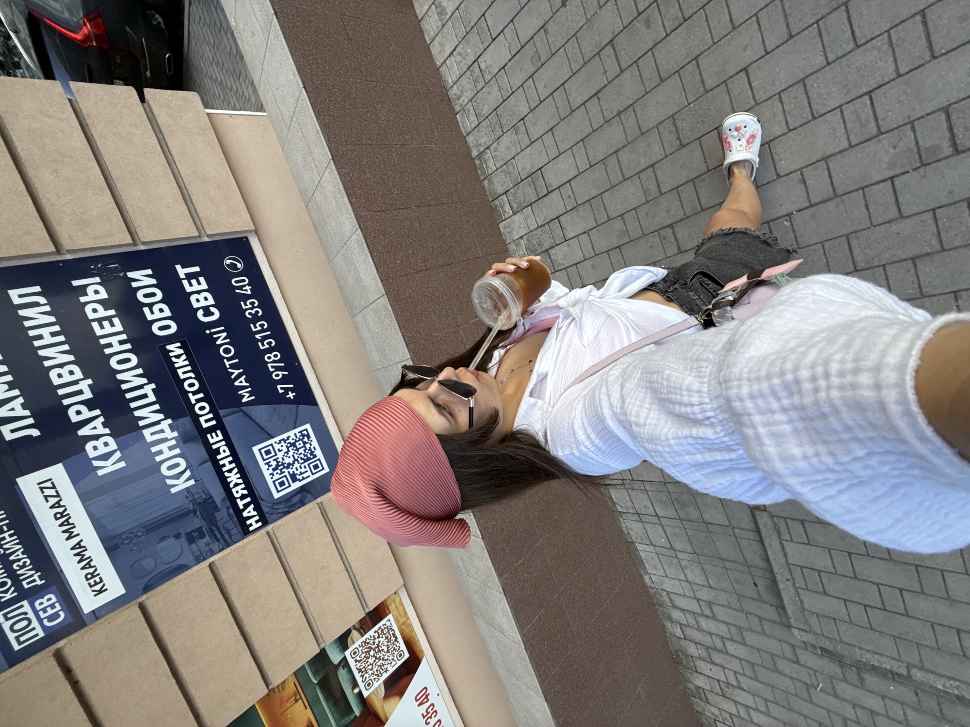 This is a selfie taken outdoors on a paved sidewalk. A young woman with dark hair, wearing sunglasses and a pink headscarf, is holding a clear plastic cup with a straw and appears to be drinking from it. She is dressed in a white shirt and a dark denim skirt. On her feet, she wears white Crocs with colorful embellishments. In the background, there is a dark-colored car.
To the left of the woman, there is a dark blue sign with white text in Russian. Visible text includes "КВАРЦВИНИЛ", "КОНДИЦИОНЕРЫ", "НАТЯЖНЫЕ ПОТОЛКИ", "ОБОИ", "MAYTONI", and a phone number "+7 978 515 35 40". There are also QR codes on the sign. The sign seems to be advertising home improvement or renovation services, such as flooring, air conditioners, stretch ceilings, and wallpaper. A portion of a cardboard box is also visible.
The scene suggests a daytime setting, possibly a sunny day given the shadows and the woman's sunglasses. The paved surface and the presence of a car indicate an urban or suburban environment. The woman's posture and the selfie-style shot convey a casual and relaxed mood.