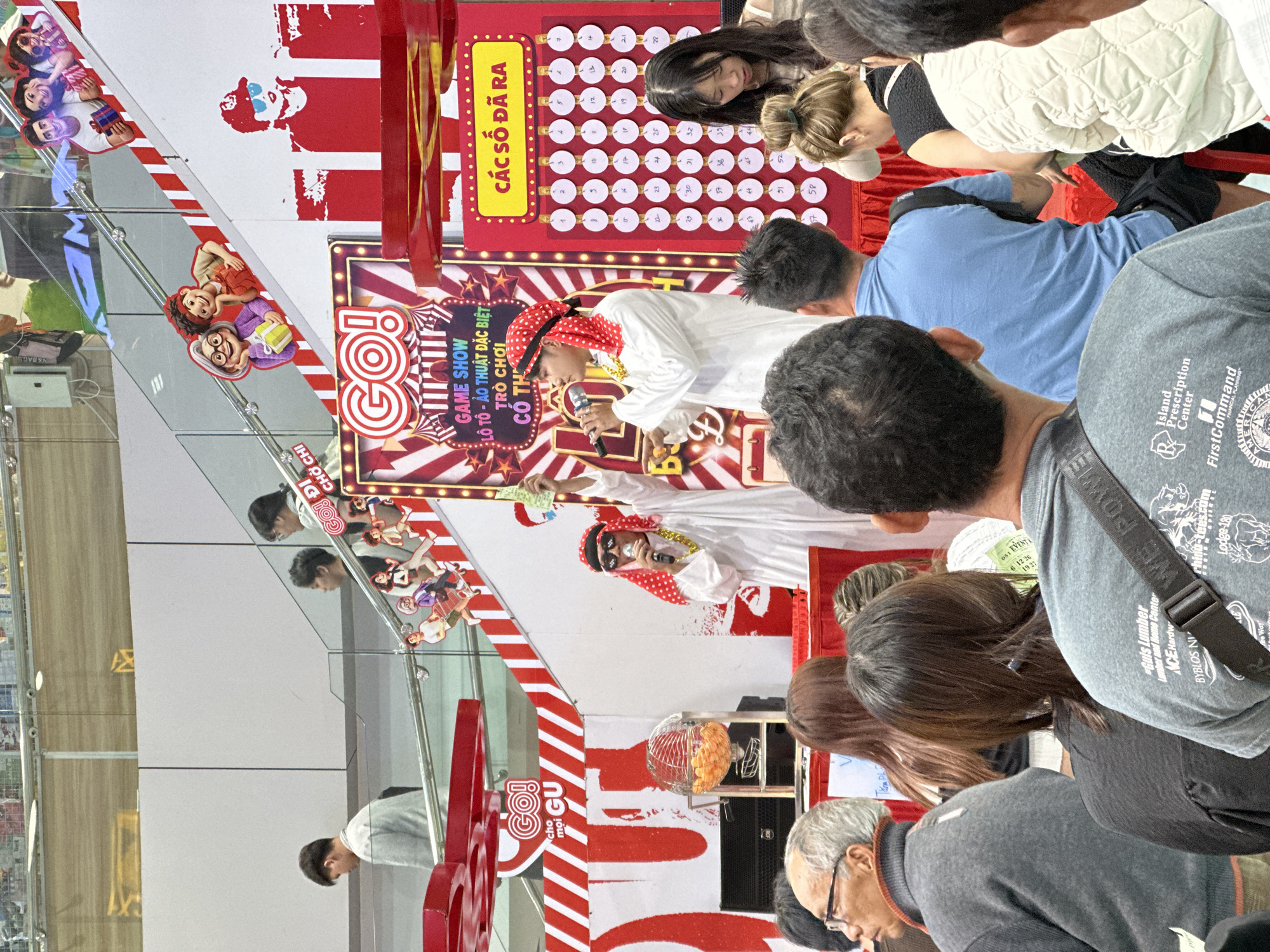 This image captures a lively scene at a game show event, likely set in a mall or public space in Da Lat, Vietnam. The central focus is a stage decorated with vibrant red and white stripes, featuring a large "GO!" logo. On stage, a man dressed in a white traditional outfit and a red-and-white checkered headscarf, complete with sunglasses and a gold chain, holds a microphone and appears to be interacting with the audience.
Behind him, a display board with numbered circles suggests a lottery or bingo-style game. The audience, a mix of men and women, are gathered around, some looking intently at the stage and others engaged in conversation. Their expressions suggest a casual and perhaps excited atmosphere.
In the foreground and scattered around the scene are various objects. A prominent red and white lollipop-shaped decoration stands to the left. A metal cage filled with orange balls, likely for the game, is visible on the stage. Several people are seen wearing casual clothing, and some have bags or accessories.
Visible text includes "GO! DI CHỜ CHI", "GO! choi GU", and "GAME SHOW LÔ TÔ - ẢO THUẬT ĐẶC BIỆT: TRÒ CHƠI CÓ TH". There are also some English words like "Island Prescription Center", "FirstCommand", and "Lumber and Home Center" on clothing and signage, indicating the presence of various businesses or brands. The overall impression is a fun and engaging public event.