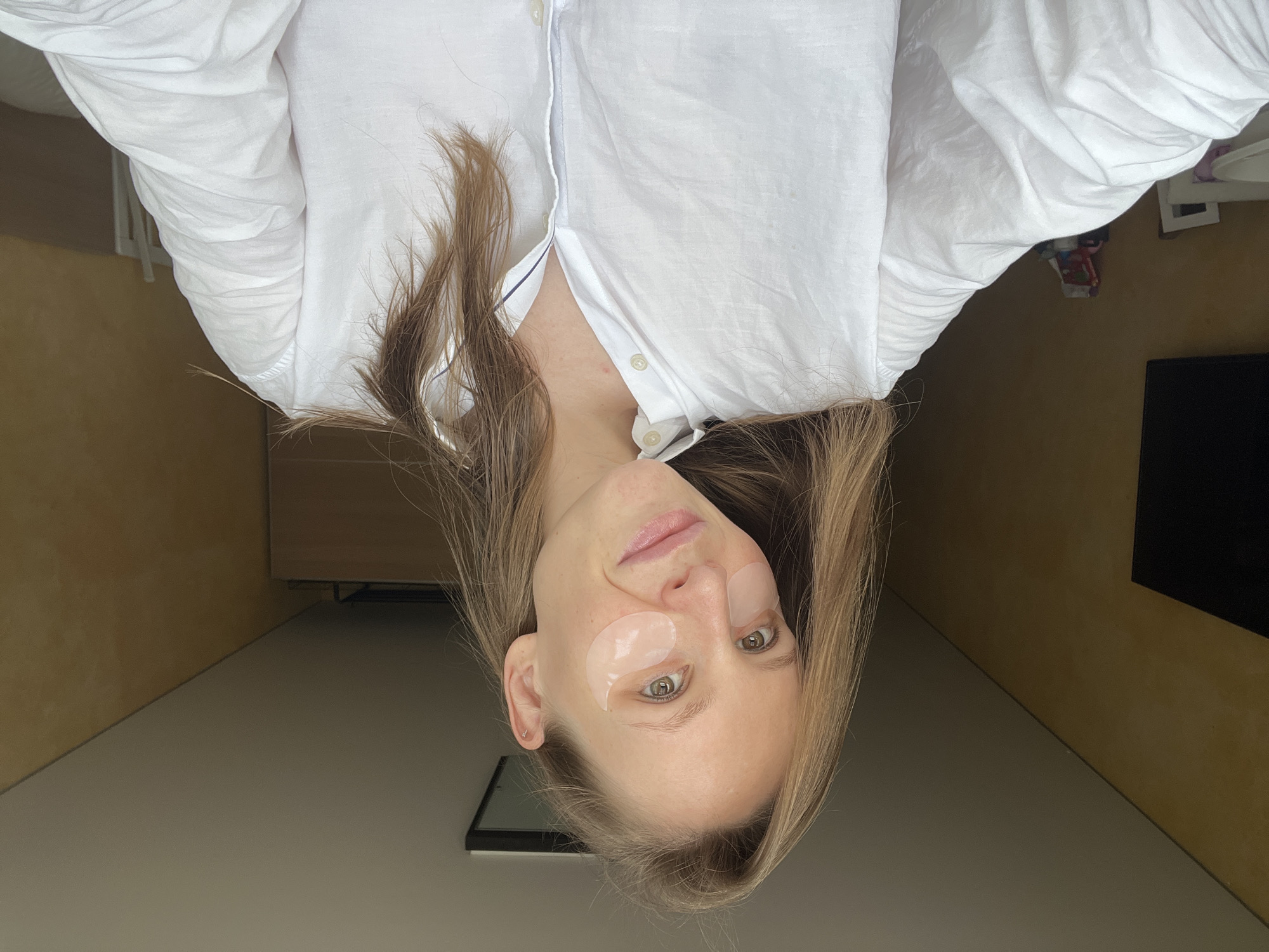 A fair-skinned woman with light brown hair wears a white button-down shirt. She has eye patches under both eyes. The background consists of a beige wall, a dark-colored television, and furniture. The image has been rotated 180 degrees. There is no information to indicate that this is in Khimki, Russia.