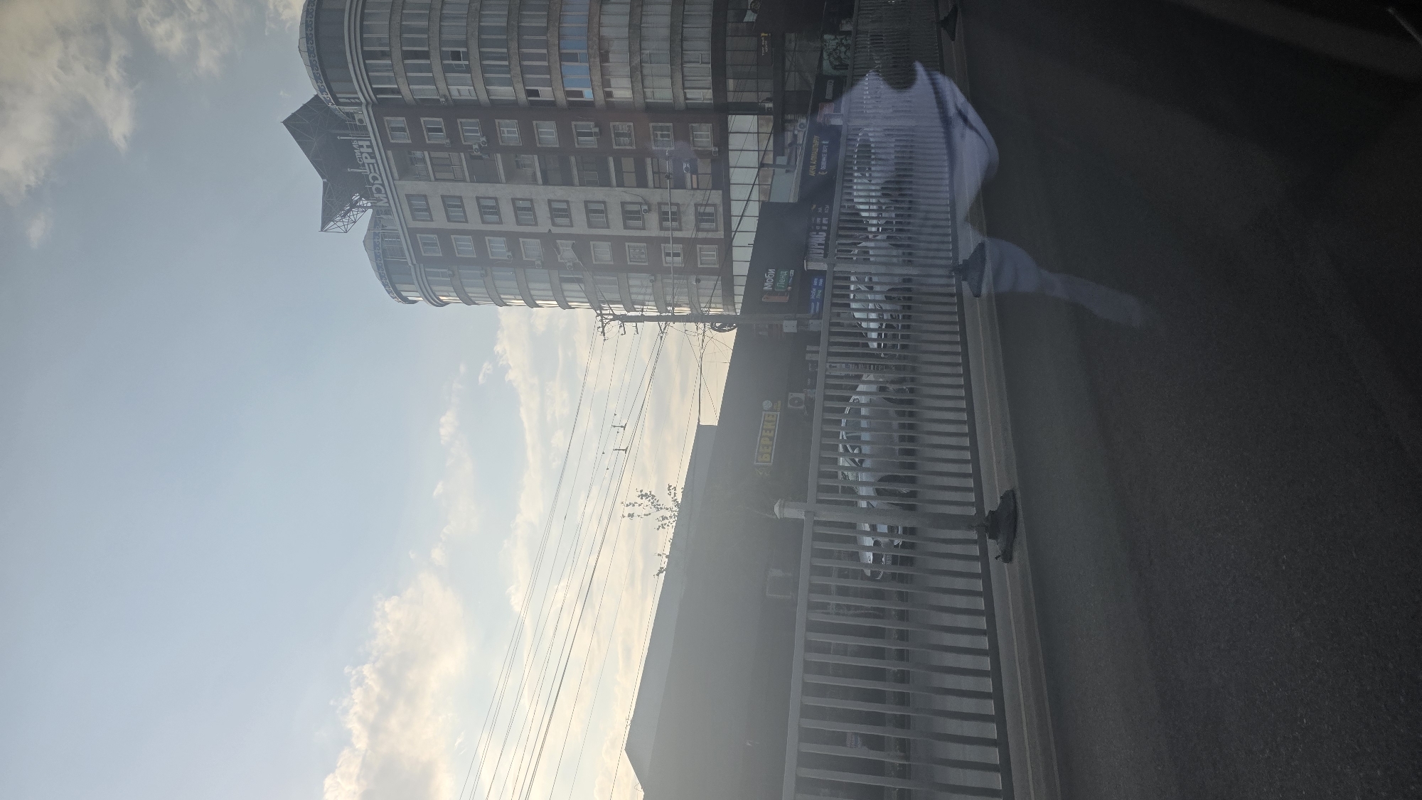 This image shows a street scene in Osh City, Kyrgyzstan, featuring a modern, cylindrical apartment building on the right side of the frame, its facade adorned with numerous windows. Adjacent to it is a lower, longer building with commercial storefronts. A prominent sign on this building displays "БЕРЕГЕ" in yellow lettering against a dark background, suggesting a business establishment. Further to the right, another sign reads "МобиЛенд" above "Новинки". A metal fence runs along the foreground, separating the street from what appears to be a parking area with several cars visible. Overhead power lines crisscross the sky, and the overall lighting suggests it is either early morning or late afternoon. The sky is a pale blue with scattered clouds.