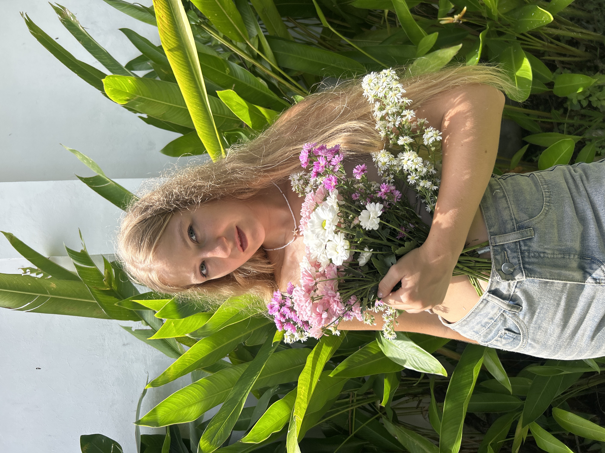 A young woman with long, fair blonde hair lies horizontally across the frame, positioned amongst lush green tropical foliage. She has fair skin, light-colored eyes, and looks directly at the camera with a neutral, calm expression. She is wearing a light-colored top and light-wash denim shorts, and a delicate beaded necklace is visible around her neck. Clutched in her hands, she holds a vibrant bouquet of small, mixed flowers, featuring shades of white, pink, and purple (magenta and lilac).
Behind her, to her left, is a textured white wall, providing a stark contrast to the abundant greenery. The large, broad leaves of tropical plants frame her, with some showing bright highlights and distinct shadows, indicating strong natural sunlight. This suggests it is daytime, likely during morning or late afternoon hours. The overall setting appears to be outdoors in a verdant, warm climate. The location is Kuta Selatan, Indonesia. There is no visible text in the image.