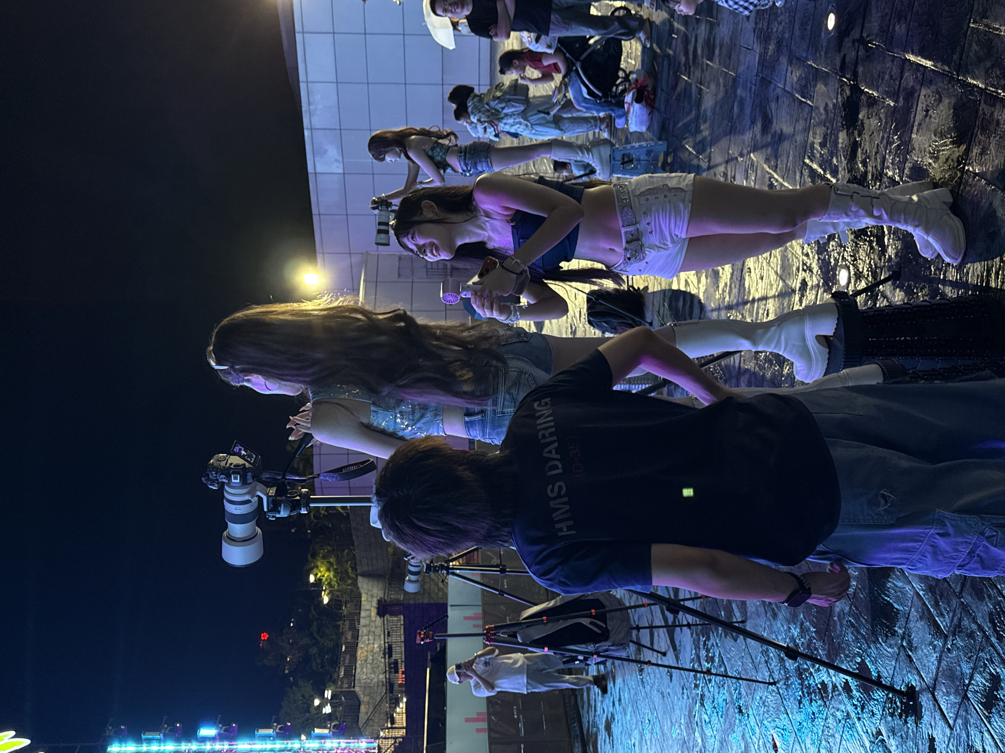 In the city of Nanshan District, China, at night, a group of young adults are gathered on a wet, paved outdoor area illuminated by stage lights and a large illuminated structure on the left. In the foreground, a young person with dark hair, wearing a black t-shirt with "HMS DARING" printed on the back and grey cargo shorts, is viewed from behind, facing a tripod with a large camera. To the right, a young woman with long brown hair, wearing a sequined blue top and white denim shorts, is standing on a stool, holding a camera and pointing it upwards. Another young woman, also with long brown hair, is in the center of the frame, wearing a dark blue crop top and white denim shorts, and white boots, holding a small handheld fan. She is looking up and to her right. In the background, several other people are visible. A man in a white t-shirt and hat is standing near a tripod, holding a camera. Other individuals are scattered throughout the scene, some with umbrellas, and a child in a stroller. The overall atmosphere appears to be a lively event, possibly a performance or gathering, with a focus on photography and entertainment.