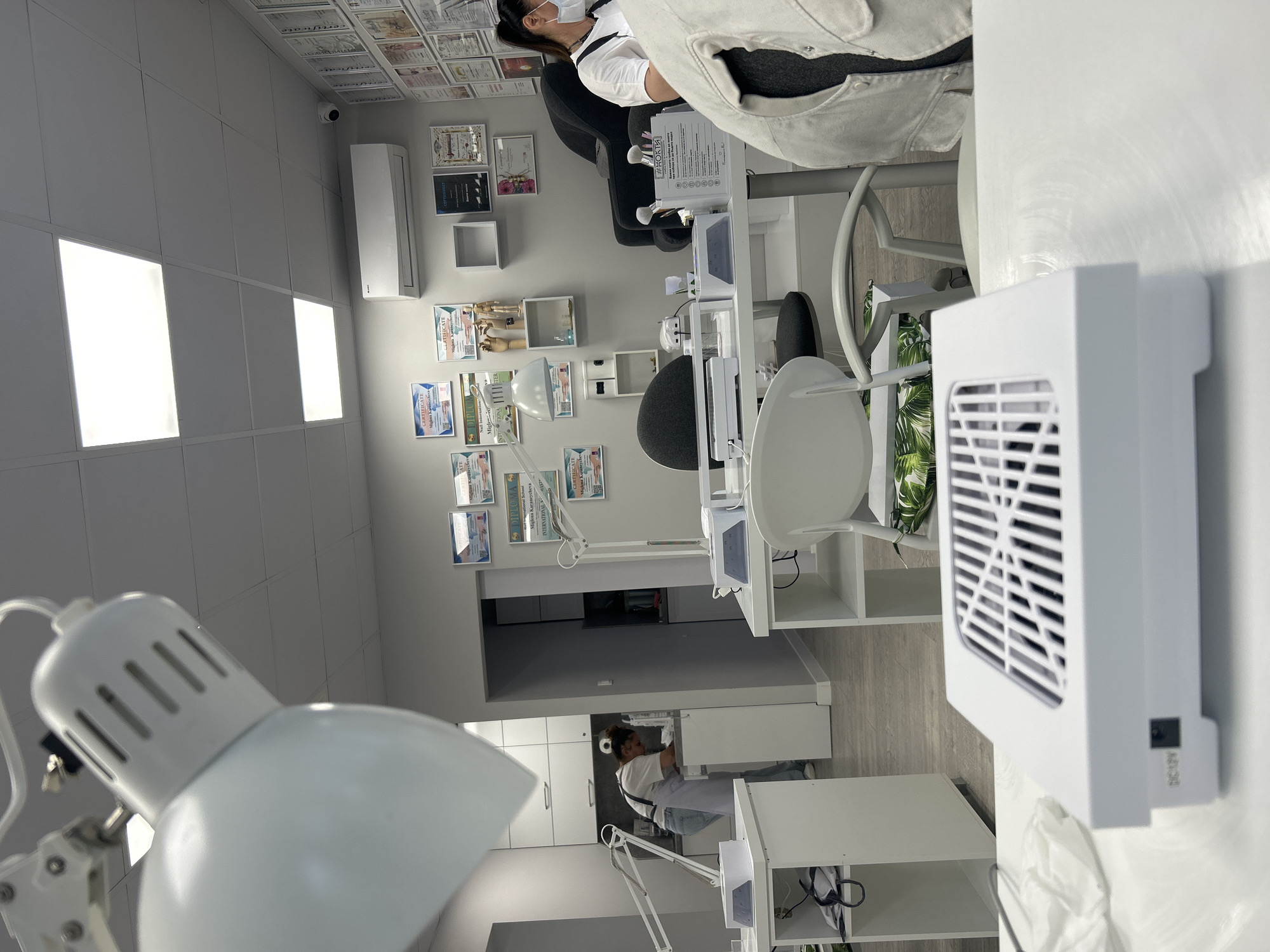 The interior of a nail salon is depicted in this shot. The room has white walls covered with framed certificates, and a drop ceiling with recessed lighting. A customer wearing a face mask is seated at a nail station in the background. There are several nail stations throughout the salon, which are furnished with white tables, chairs, and lamps. A white fan for removing dust from nail filing is on one of the tables. The camera is close to a white lamp with a gooseneck. The scene is well-lit.