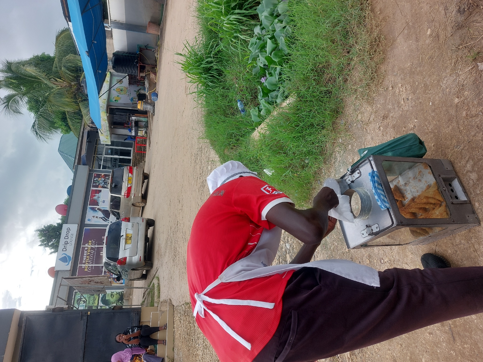 The image shows a street food vendor in what appears to be Dar es-Salaam, Tanzania. The vendor, wearing a red shirt, white apron, and chef's hat, is preparing food from a metal and glass display case. Inside the case, there are several pastries. He is placing the food into a paper container. The backdrop includes a street scene with parked cars, a storefront labeled "Drip Drop LOUNGE," and other business signage. Vegetation grows along the side of the dirt road where the vendor is positioned. Other people are sitting on the steps of the building behind a black iron gate. The overall environment suggests a busy, urban area with both commercial establishments and street vending activity. The OCR suggests that the location is Dar es Salaam, Tanzania.