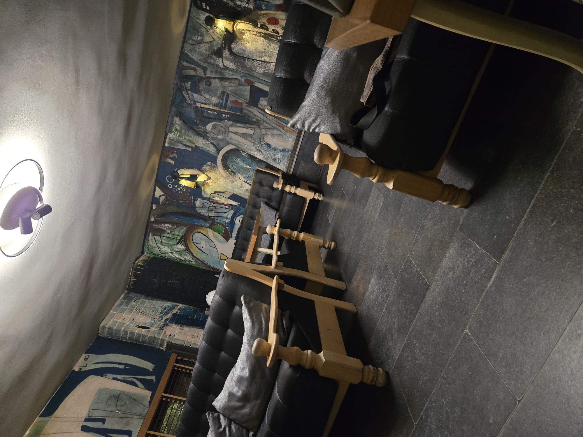 The image captures an interior room, presented at a tilted angle, showcasing what appears to be a lounge or common area. A prominent feature of the space is a large, colorful mural covering the walls, characterized by abstract and stylized human-like figures or forms in various shades of blue, white, gray, and hints of red and green. The artwork gives the room a unique and artistic ambiance.
In the foreground and arranged along the mural are several dark-colored sofas or benches, upholstered in a material that appears to be black leather or a similar dark fabric. Some gray throw pillows are placed on these seating arrangements. Interspersed between and in front of the sofas are multiple light-colored wooden structures, possibly parts of tables or decorative balusters, featuring turned legs and a smooth finish.
Artificial lighting is evident, with a bright, wall-mounted fixture with a purple element visible on the left side of the frame, casting illumination on the light-colored upper wall or ceiling. Other light sources within the scene highlight portions of the mural. The floor is composed of large, dark gray or charcoal-colored rectangular tiles, which exhibit a somewhat textured or speckled appearance.
No people are visible in the image, and there is no specific activity taking place, suggesting the room is currently unoccupied. The reliance on artificial lighting implies that it could be evening or night. The overall impression is one of a modern or artistically decorated interior designed for relaxation or gathering. No discernible text is visible within the frame.