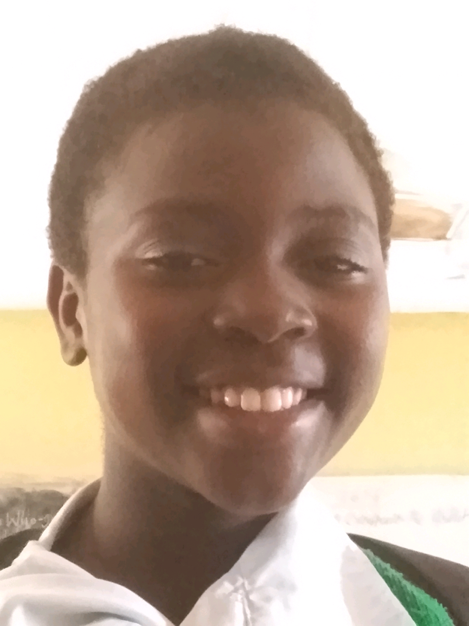 A close-up, head and shoulders shot shows a young girl smiling widely at the camera. She has dark skin and short, dark, curly hair. She is wearing a white collared shirt, and a glimpse of a green garment is visible beneath it. The background is a blurred mix of yellow and white. The image appears to have been taken indoors, possibly in a classroom or home setting, with a light source illuminating the subject from the front. The overall impression is one of warmth and joy.