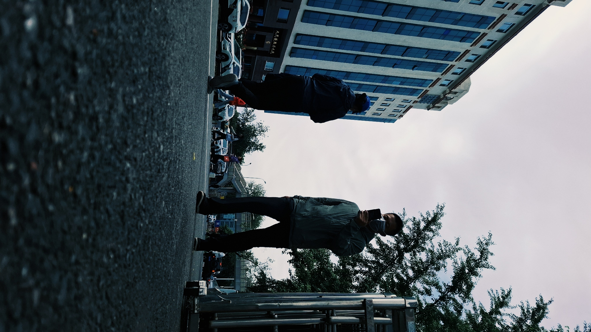 A scene in Dongcheng District, China, shows two people on a street. The perspective is rotated approximately 90 degrees clockwise. The foreground features a person standing on a sidewalk, facing right, wearing dark pants and shoes, a dark jacket, and a light-colored mask. This individual is holding a dark object, possibly a phone. Behind them, there are trees and a metal structure. Cars and a building line the street. Above the street, another person, dressed in dark clothing and a blue hat, appears to be doing push-ups in the street, with cars and a building behind them. The sky is overcast. The ground is paved.