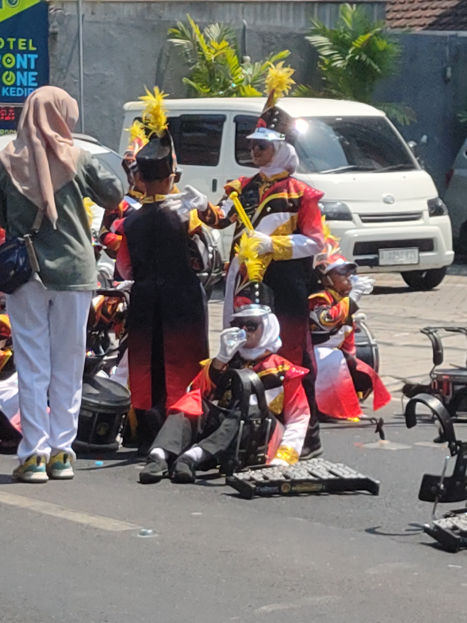 In Ngasem, Indonesia, a group of children in marching band uniforms is gathered on a street. The uniforms are colorful, featuring red, gold, and black accents, and include tall, yellow plumed hats. Some children are holding musical instruments. One child is drinking from a bottle. In the background, a white van is parked on the side of the road, and a sign for "HOTEL MONT ONE KEDIRI" is visible. The scene appears to be during a sunny day, with palm trees and buildings forming the backdrop.