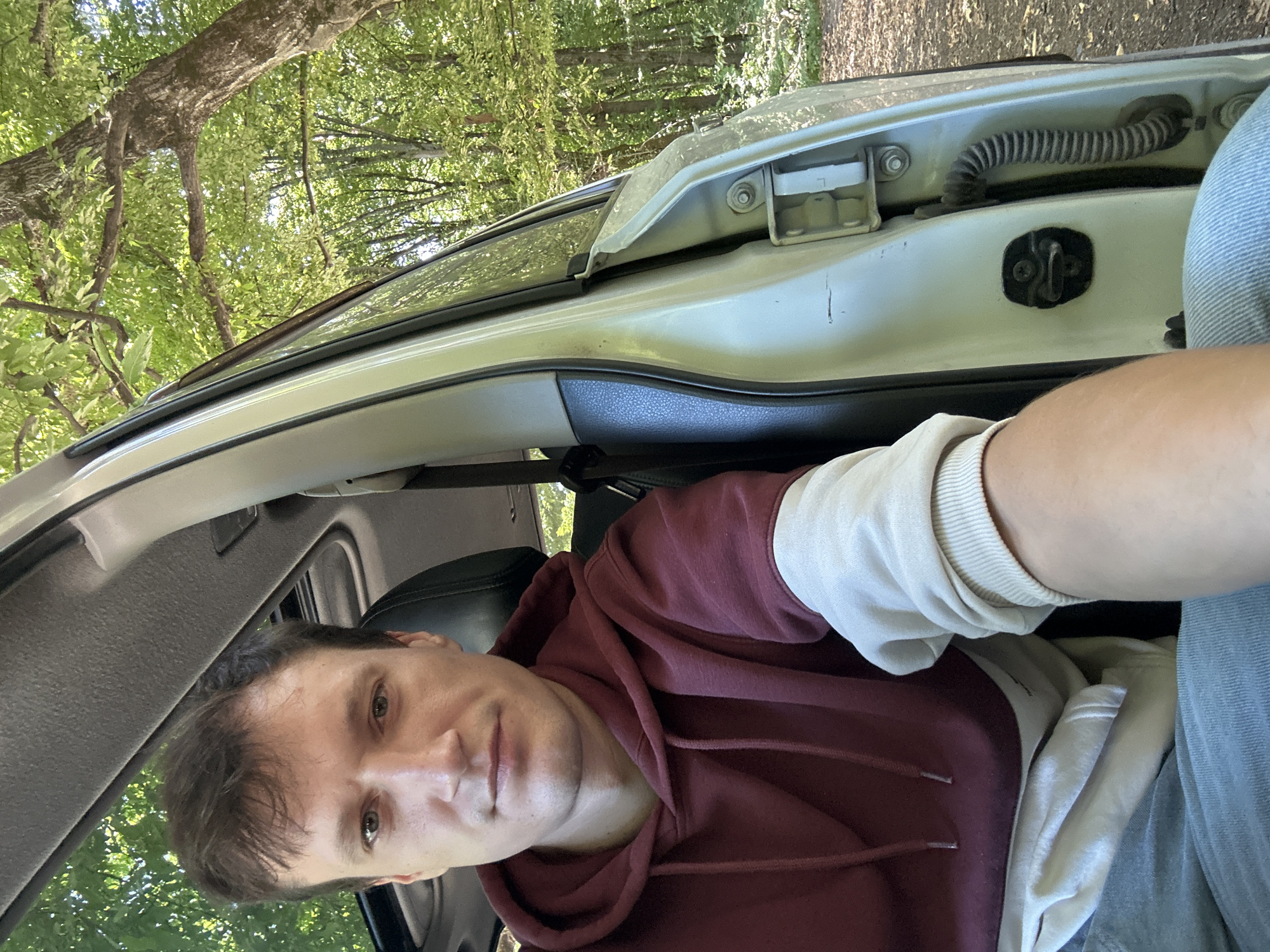 A young man is seated in the driver's seat of a car, with the passenger door open. He is taking a selfie, looking directly at the camera with a neutral expression. He is wearing a maroon and cream hoodie and blue jeans. The car is parked on a dirt road surrounded by a dense forest with lush green foliage. Sunlight filters through the trees, casting dappled shadows on the ground and the car. The car's interior is visible, including the dashboard and the sunroof. The background shows the trees and a pathway leading into the woods. The image appears to have been taken during daytime.
