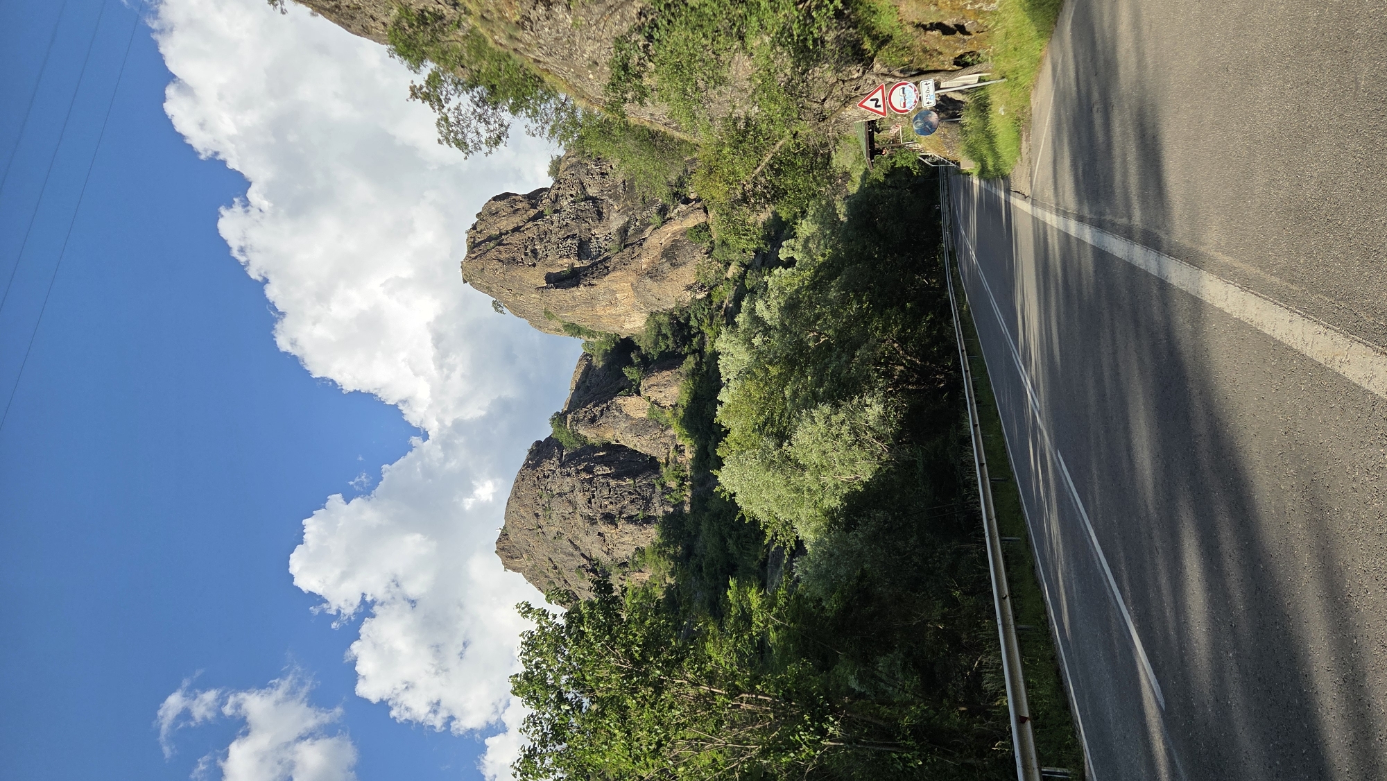 The image depicts a scenic mountain road in the vicinity of Rila, Bulgaria. The road, paved and marked with white lines, curves gently to the right, bordered by a metal guardrail on the left and lush green foliage on the right. Towering rock formations, weathered and rugged, dominate the background, with trees and shrubs clinging to their slopes. The sky is a vibrant blue, adorned with fluffy white clouds. Several road signs are visible on the right side of the road, indicating a sharp curve ahead and potentially other traffic regulations. The overall impression is one of a tranquil, natural landscape, likely a popular route for scenic drives or outdoor excursions in the Rila region.