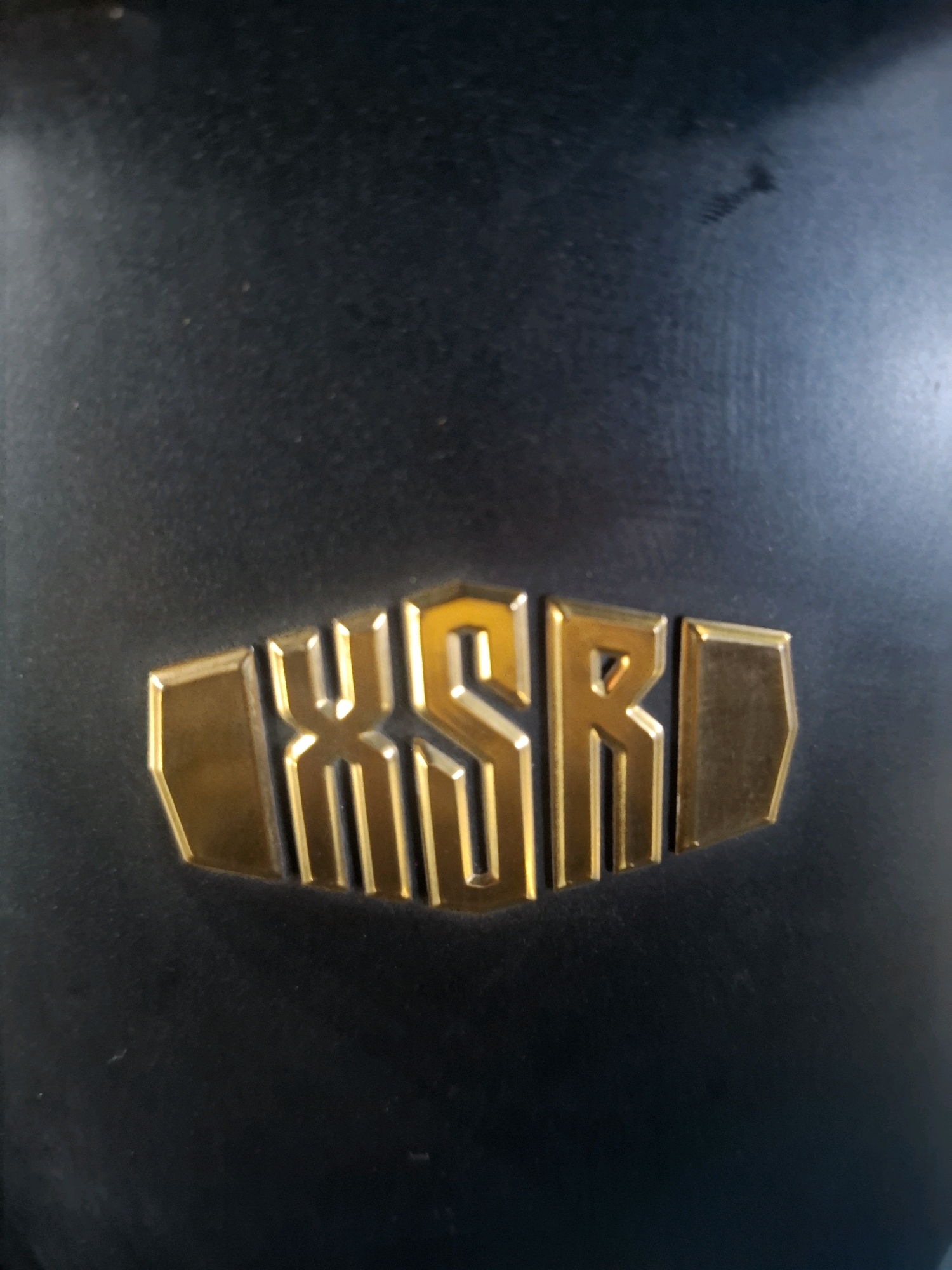 A close-up, slightly angled, full-frame shot captures a metallic, golden emblem against a dark, textured background. The emblem is shaped like a horizontal rectangle with beveled edges and contains the stylized letters "WSR" in the center. The "W" is formed by two vertical lines connected by a V-shape, the "S" is a classic cursive S, and the "R" has a vertical stem with a loop and a diagonal leg. The letters are outlined and filled with a bright gold, with subtle reflections of light on their surfaces. The background appears to be a dark, matte material, possibly fabric or a coated surface, with some minor scuffs or imperfections visible. The overall lighting suggests an indoor environment with a direct light source illuminating the emblem from the front. The specific location within Perumahan Puri Indah, Indonesia, cannot be determined from the provided image alone, as it focuses solely on the emblem and its immediate surroundings.