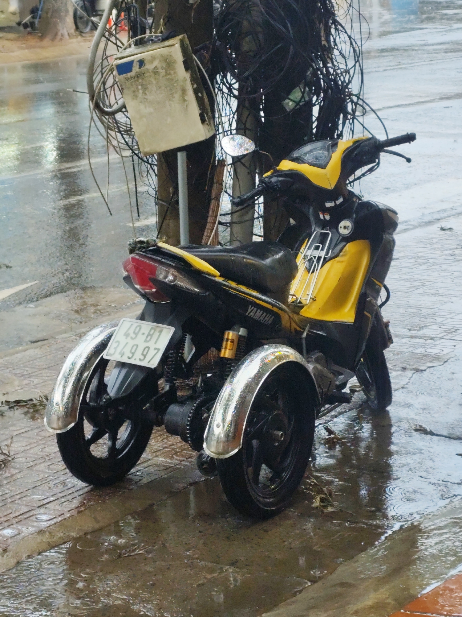 A yellow and black Yamaha scooter with three wheels is parked on a wet sidewalk next to a telephone pole with tangled wires in Lang Biang - Đà Lạt, Vietnam. The scooter's license plate reads "49-B1 349.97". The sidewalk is wet and reflects the light. The road behind the scooter is also wet, indicating it has been raining.