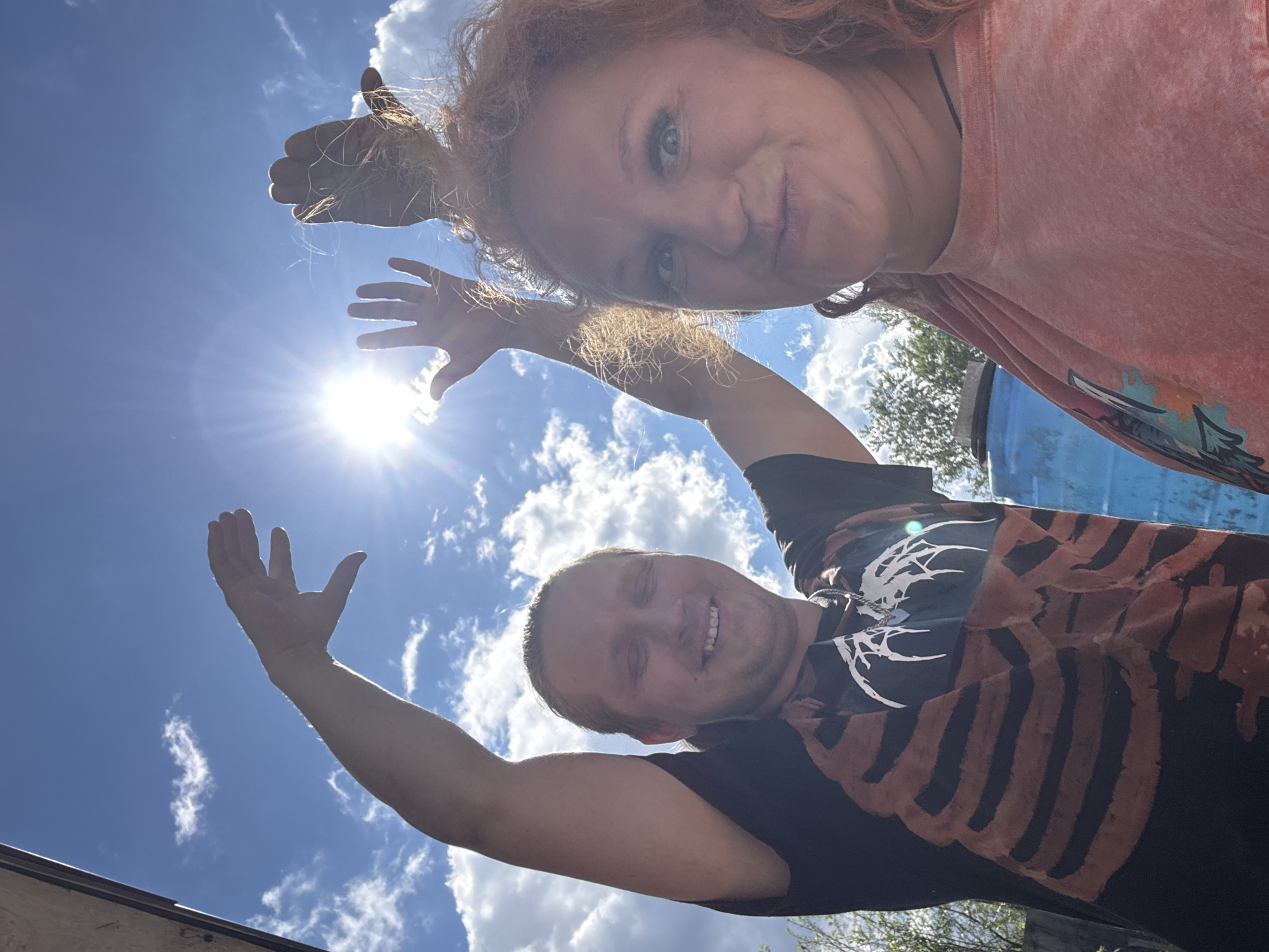 This vertical image captures two individuals, a woman and a man, posing outdoors in Chaikovsky, Russia, on a bright, sunny day. The photograph is taken from a low angle, looking upwards towards the sky, creating a playful perspective.
In the upper half of the frame, a woman with reddish-blonde curly hair and light eyes looks down at the camera with a playful expression. She is wearing a light orange t-shirt with a graphic design. Her hands are raised towards the sun, appearing partially silhouetted against the bright light.
Below her, an adult man with light brown hair smiles broadly at the camera. He is wearing a dark t-shirt featuring a prominent light-colored skeleton or ribcage design across the chest. His arms are also raised towards the sky, mirroring the woman's pose, and are silhouetted by the intense sunlight.
The sun is a dominant feature in the center-left of the image, casting a strong glare and lens flare. The background consists of a clear blue sky dotted with scattered white clouds, indicating fine weather during what appears to be midday or early afternoon. Some green tree foliage is visible in the far background, particularly on the right side, along with a partially obscured blue object, possibly a barrel or container. The overall mood conveyed by the subjects' expressions and the bright setting is cheerful and relaxed. There is no visible text in the image.