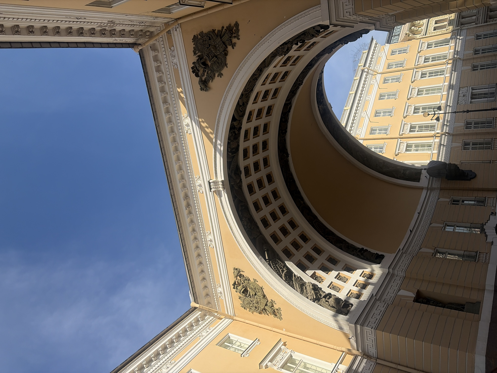 The image shows a low-angle view of a large architectural archway against a clear blue sky. The archway is made of yellow stucco with white decorative trim and ornate details, including what appears to be a double-headed eagle emblem on the left side of the arch. The interior of the arch is dark, revealing a coffered ceiling with square-shaped recesses. Through the arch, another yellow building with many windows and white trim is visible. The scene suggests an urban environment with classical architecture.