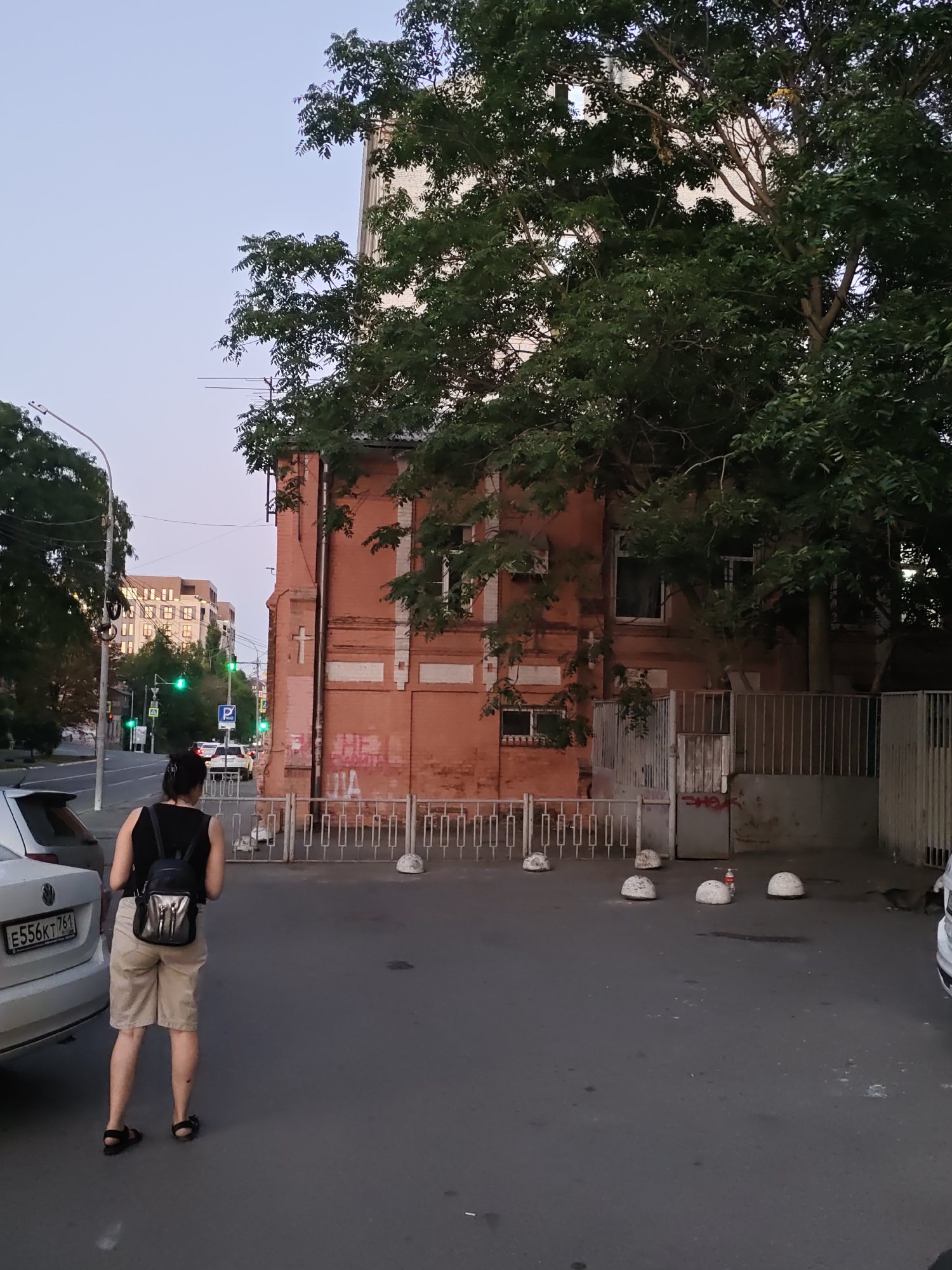 The image depicts an outdoor scene on a street in Rostov-on-Don, Russia, likely during dusk or early morning given the lighting. In the foreground, a woman with dark hair, wearing a black tank top, beige shorts, and sandals, stands facing away from the camera. She is carrying a small, metallic-colored backpack. To her left, the rear of a white car with a license plate reading "E556KT 761" is visible.
The background features a brick building with some graffiti on its lower section. A white metal fence with a decorative pattern runs along the front of the property. Several white, dome-shaped bollards are placed in front of the fence. To the left of the brick building, a street is visible with cars and traffic lights. A modern, multi-story building with many windows can be seen in the distance. Trees are present throughout the scene, with dense foliage on the right side of the brick building. The sky is a pale blue, suggesting low light conditions.