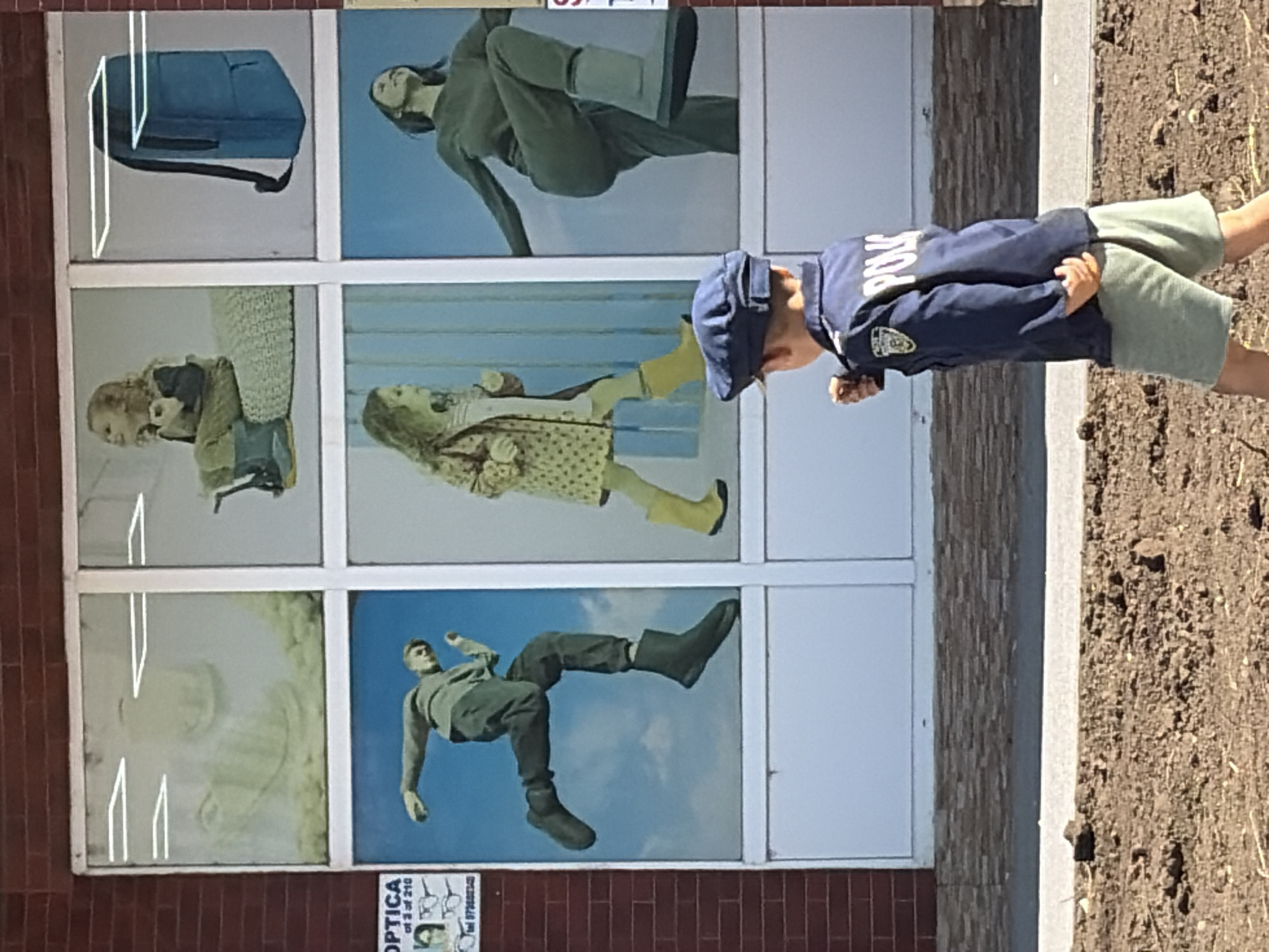 A young boy dressed as a police officer walks in front of a storefront in Criuleni, Moldova. The storefront has a large display window divided into six panels. The top row of panels features a hazy image of a face, a young girl sitting on a cushion with boots, and a blue backpack. The bottom row shows a man in mid-stride, a young girl in a patterned dress and yellow boots, and a woman in a green outfit with boots. The boy in the foreground wears a blue police uniform with "POLICE" written on the back and green shorts. He is walking to the right, looking towards the storefront. The ground in front of the store is a mix of dirt and gravel. The building has a red brick facade above the display windows.