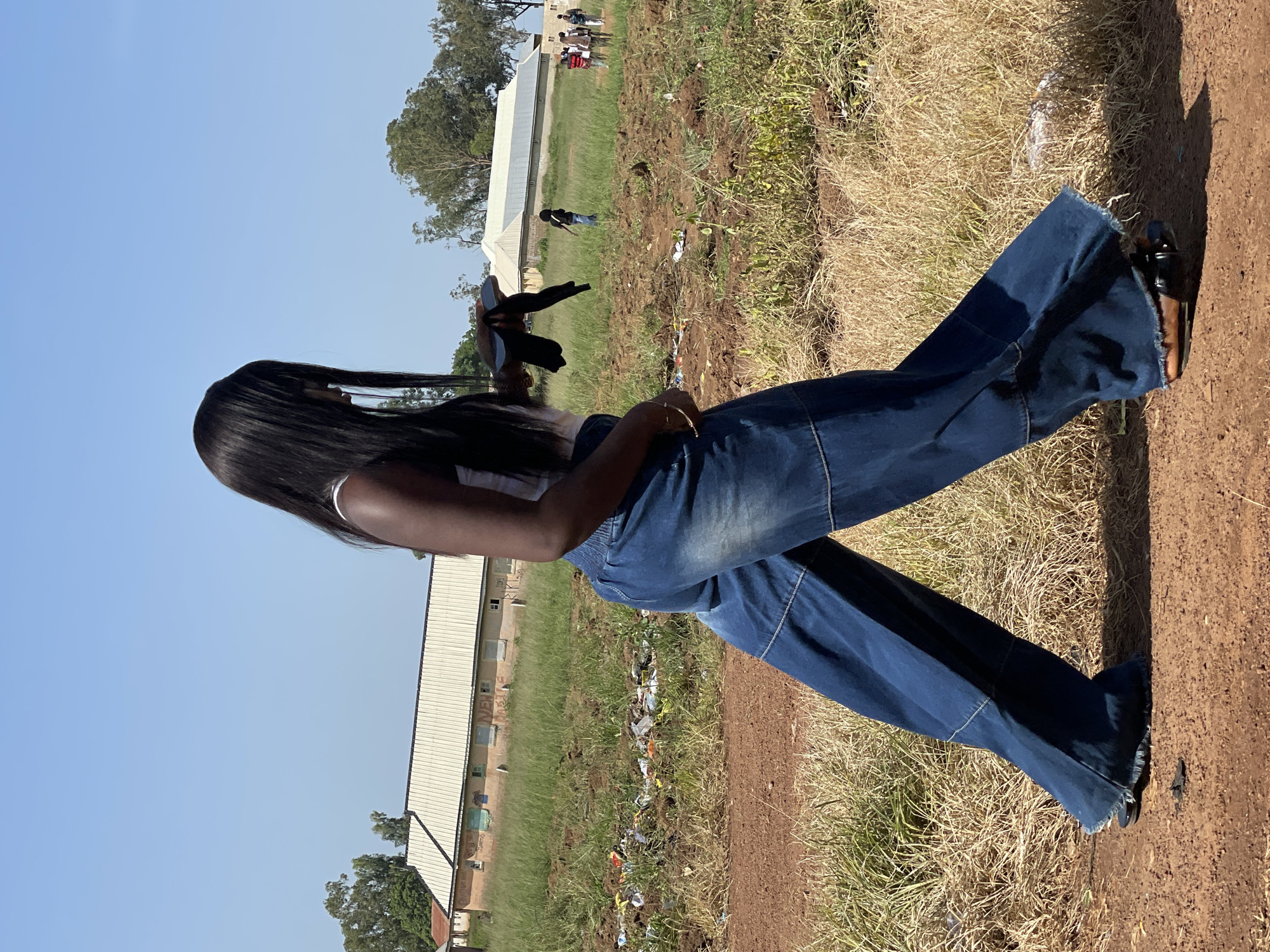 A young Black woman with long, dark hair is walking towards the right side of the frame in a field. She is wearing a white tank top and wide-legged dark denim pants. Her right hand is holding a dark object. In the background, there is a long, light-colored building with a metal roof. Further in the distance, several other people are visible, some standing and some walking. The ground is a mix of dry grass, dirt, and some scattered debris. The sky is clear and blue. The scene appears to be in an outdoor, rural or semi-rural area.