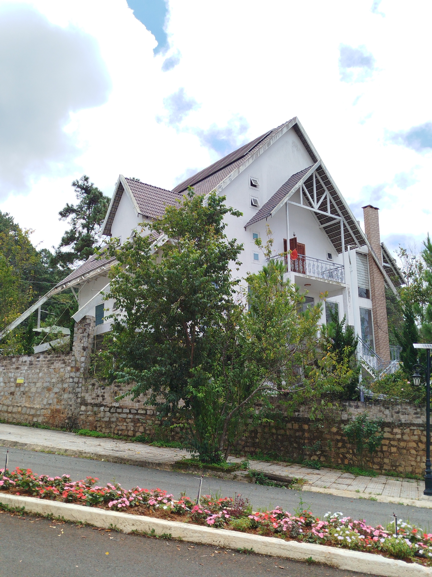 The image shows a multi-story white house with a brown tiled roof, featuring a prominent chimney made of light brown bricks. The house has a balcony with an ornate railing and a red object visible on it. There are several windows, some of which are open. The house is situated behind a low stone wall and surrounded by lush green trees. In the foreground, a paved road is visible with a strip of colorful flowers, predominantly pink and red, planted along its edge. A lamppost stands on the right side of the image. The sky is overcast with some patches of blue. The location is identified as Lạc Dương, Vietnam.
