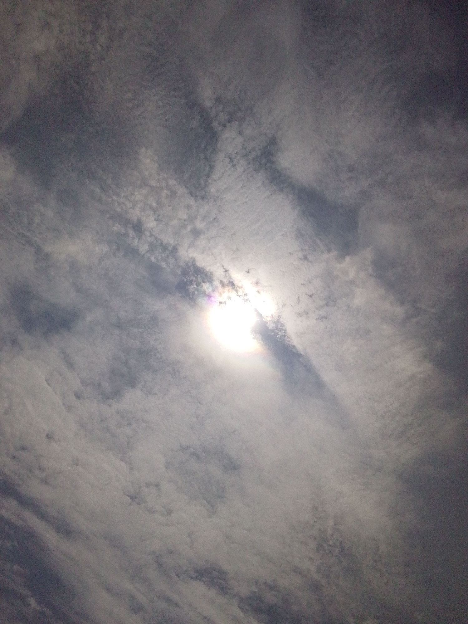 This image shows a cloudy sky with the sun partially obscured by clouds. The sun appears as a bright, white orb with a subtle rainbow-like halo around it. The clouds are varied in texture and density, with some appearing thin and wispy, while others are thicker and more opaque. The overall impression is of a hazy or overcast day, with the sun's light diffused through the atmospheric conditions. There are no people, subjects, or objects other than the natural elements of the sky and clouds. The location in Serti, Nigeria, is noted as a contextual detail and does not influence the visual description of the image itself.