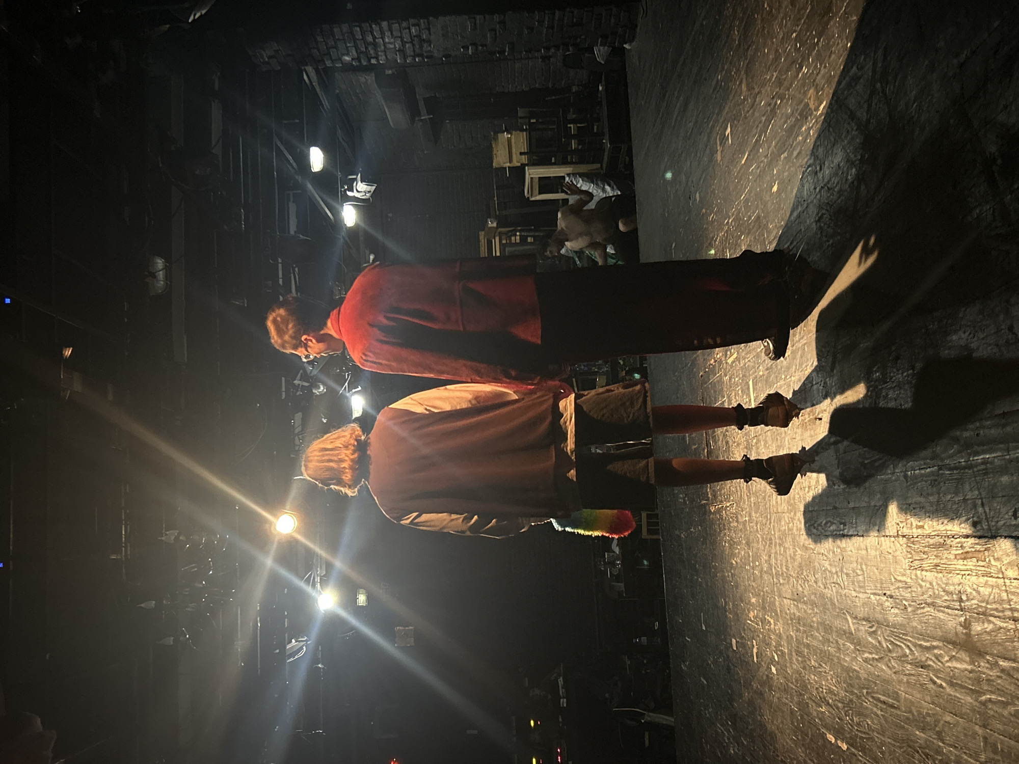 Two individuals stand on a wooden stage, facing away from the camera. The individual on the left has shoulder-length blonde hair and is wearing a maroon jacket over a light-colored shirt and dark shorts. They are holding a rainbow-colored fluffy object. The individual on the right is wearing a red jacket and dark pants. The stage is lit by bright spotlights from above, casting long shadows. In the background, there are brick walls, wooden crates, and other stage equipment. The overall environment appears to be an indoor theater or performance space.