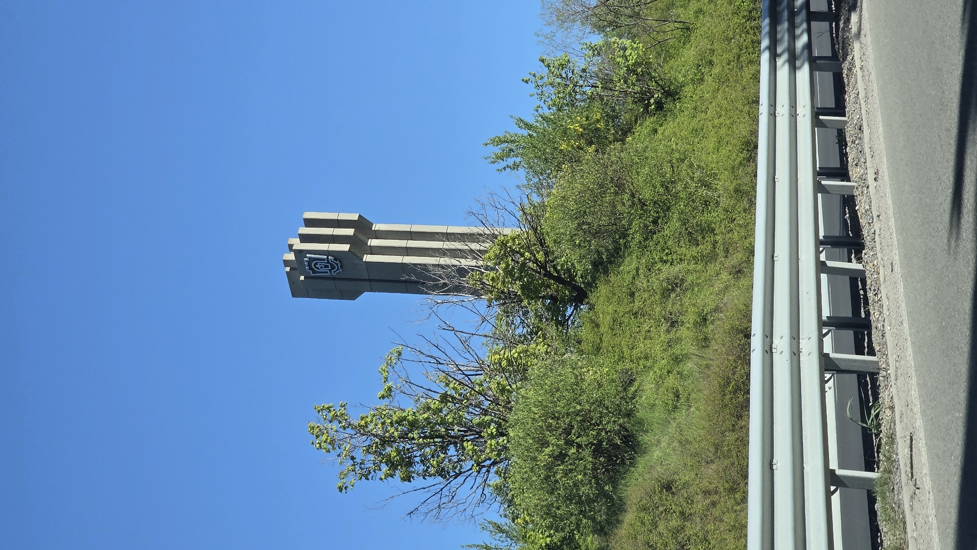 The image depicts a tall, light-colored concrete tower under a clear blue sky. The tower has a layered, blocky design at the top and is adorned with a circular emblem. The tower is located in Marchaevo, Bulgaria. To the right of the tower, a hill covered with lush green vegetation and trees is visible. A metal guardrail runs parallel to the edge of a road or highway, partially obscuring the bottom right corner of the image. The photograph appears to have been taken from a moving vehicle, likely traveling along the highway adjacent to the tower and the hillside.