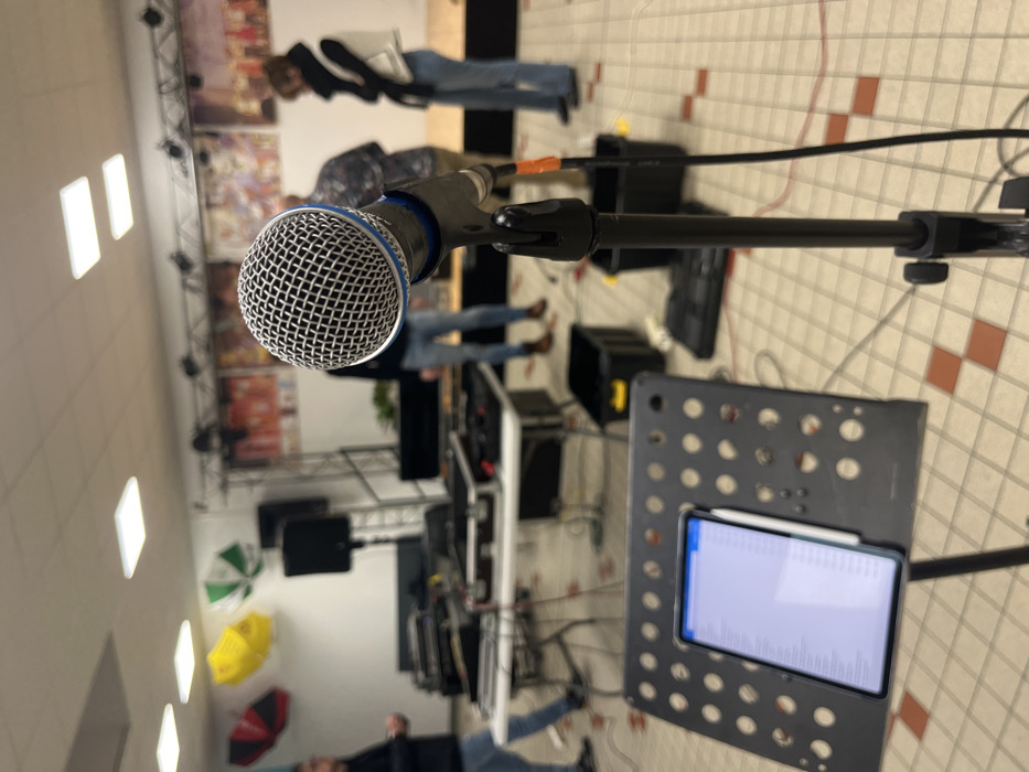 This image depicts an indoor event space, likely a multi-purpose hall, located in Coudekerque-Branche, France, during what appears to be an equipment setup or rehearsal.
In the immediate foreground, a dynamic microphone with a silver grille and blue band is mounted on a black stand, positioned centrally. To the left of the microphone stand, a black perforated music stand holds a tablet displaying a scrollable list, possibly lyrics or a setlist, with a white stylus attached to its side.
The midground is largely out of focus, showing multiple individuals and professional audio equipment. At least four people are visible: one on the far left wearing dark clothing, two indistinct figures in the center-right, and one on the far right wearing a white vest over dark clothing and blue jeans. Two white rectangular tables are laden with audio mixing consoles, cables, and various electronic devices. Two black speakers are visible on stands behind these tables. Several black plastic crates or containers are on the floor.
The background reveals a low stage or platform draped in black, positioned against a white wall. The wall displays three open umbrellas as decorative elements: one solid yellow, and two striped green and white. Further along the wall, several pieces of framed artwork or posters are visible above the stage. The ceiling is white with multiple rectangular fluorescent light fixtures. The floor is covered with light-colored tiles featuring a grid pattern of alternating light beige and reddish-brown squares. The overall scene indicates preparation for a performance or public event.