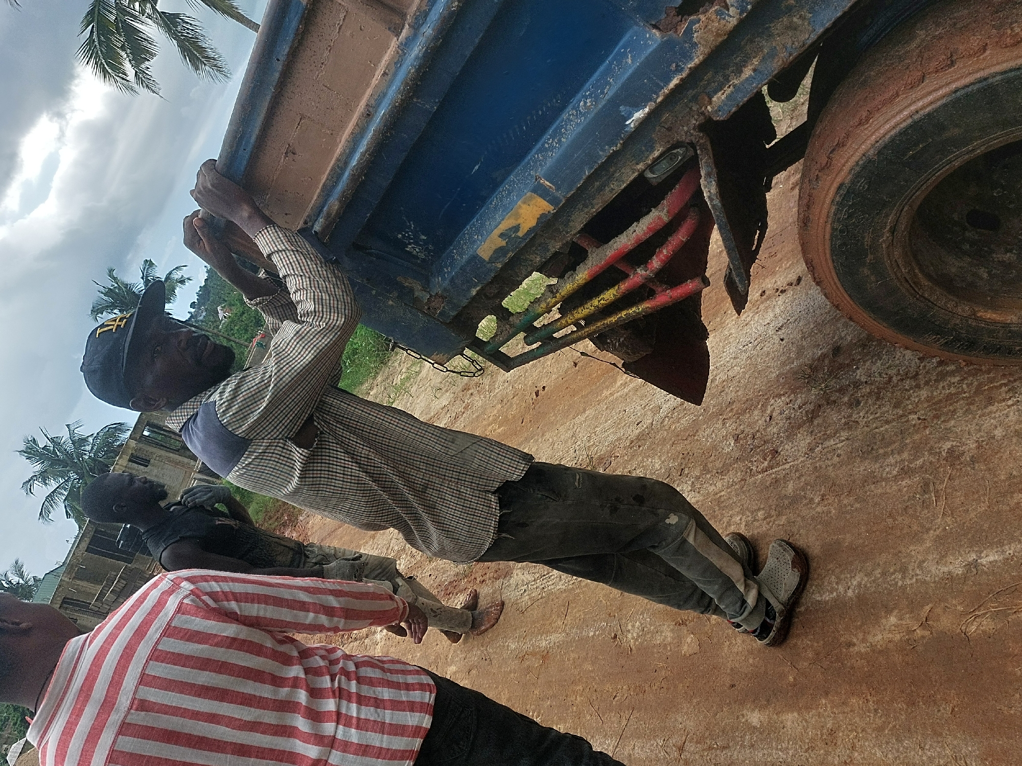 A medium shot captures three men gathered around an old, rusty blue truck on a reddish-brown dirt road. The central figure, a man wearing a plaid shirt, dark pants, and a black cap with yellow "Tf" lettering, holds onto the side of the truck bed. He is looking towards the camera with a neutral expression. His grey athletic-style shoes are visible, marked with dirt. In the foreground, a man in a red and white striped shirt is seen from behind, and another man in a dark shirt is partially visible behind the central figure.
The truck's bed is blue but heavily rusted, with the undercarriage showing yellow and red painted metal bars also corroded. A large, mud-covered tire is visible on the right. The ground appears muddy in places, suggesting recent wet conditions. In the background, palm trees and lush green foliage indicate a tropical environment. A partially constructed concrete building is also visible behind the men. The sky is overcast with dense clouds, suggesting daytime with diffused light. No other discernible text is visible besides the "Tf" on the cap. The overall scene depicts a moment of manual activity or a break from work in an outdoor setting.