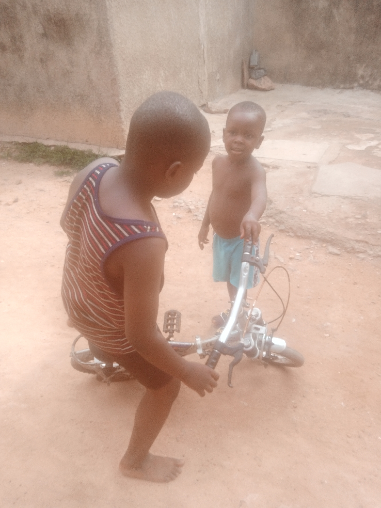 Two young children are outdoors on a dusty ground. The child on the left, facing away from the camera, is wearing a red and white striped tank top. They are kneeling beside a silver bicycle, holding its handlebars. The child on the right, facing the camera, is shirtless and wearing light blue shorts. They are standing and appear to be interacting with the child on the left and the bicycle. The background consists of a textured wall and more dusty ground.