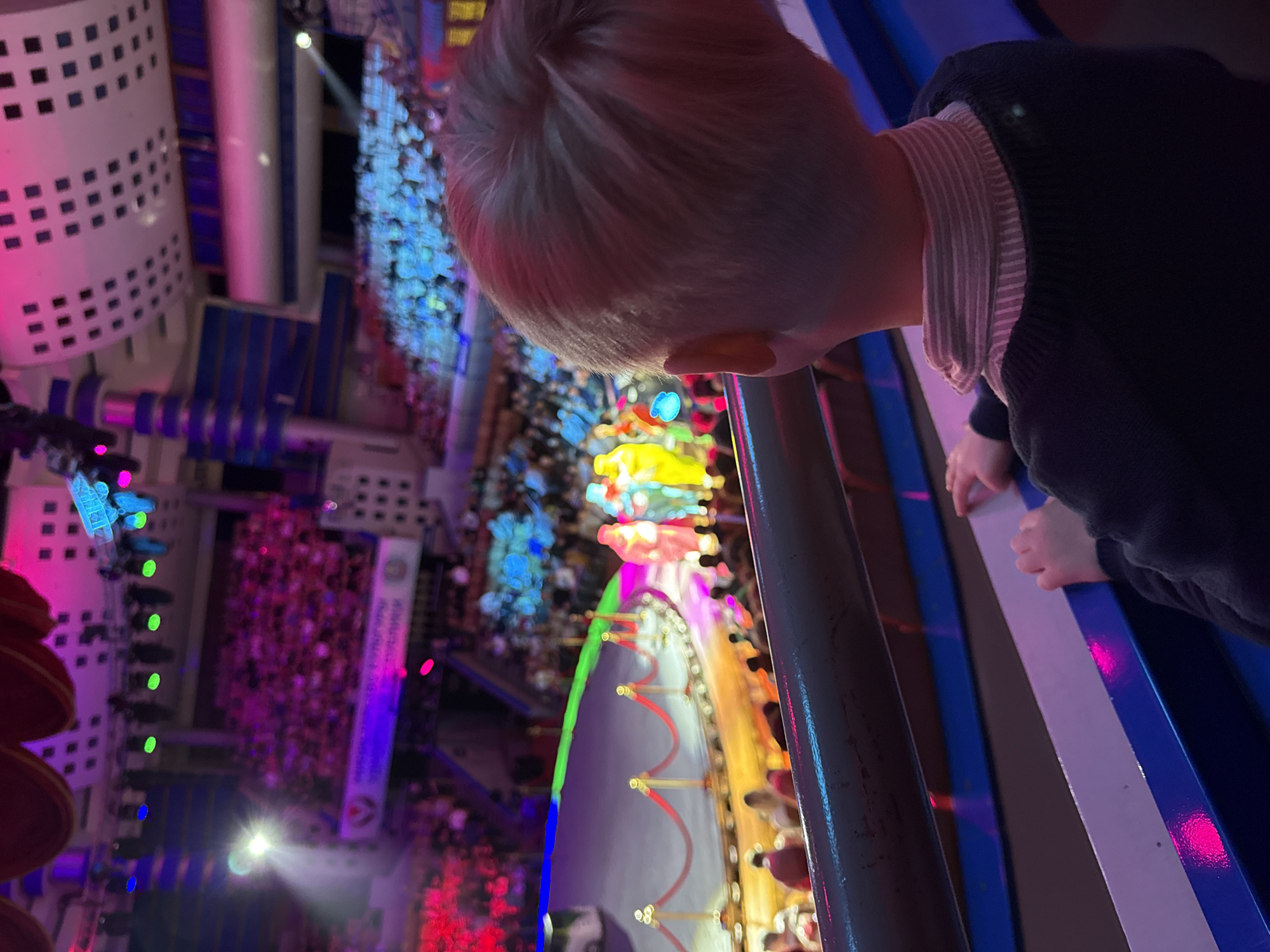 A low-angle, tilted indoor shot shows a child looking over a barrier at a colorful performance or event in a large venue. The child, with short blonde hair and wearing a dark sweater, is leaning over a metal and blue striped barrier. The background features a complex arrangement of lights, structures, and what appears to be a large crowd of people. Dominant colors include pink, blue, green, and yellow, suggesting a dynamic and visually stimulating environment. Given the context of Izhevsk, Russia, this may be a view from a higher level in the State Circus of Udmurtia.
