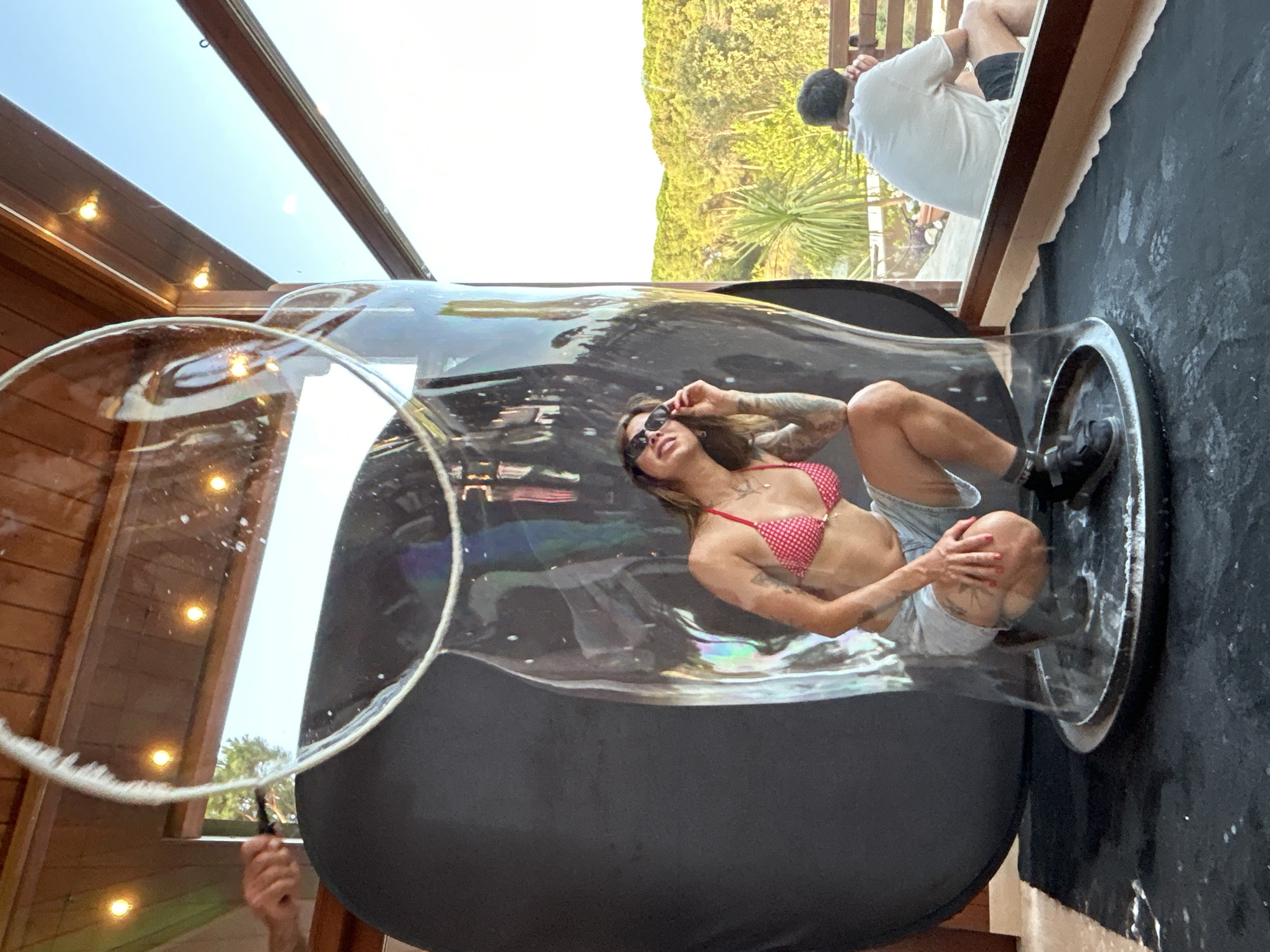 A woman in a red and white polka dot bikini top and grey shorts is inside a large, transparent bubble. She is crouching down and adjusting her sunglasses. The bubble is placed on a black mat. In the background, through a large window, a man in a white t-shirt and black shorts is sitting on a wooden deck, looking out at a lush green landscape with trees. The setting appears to be outdoors, possibly a resort or a private residence, given the architecture of the wooden ceiling and the view of the vegetation. The location is Sant Vicenç De Montalt, Spain.