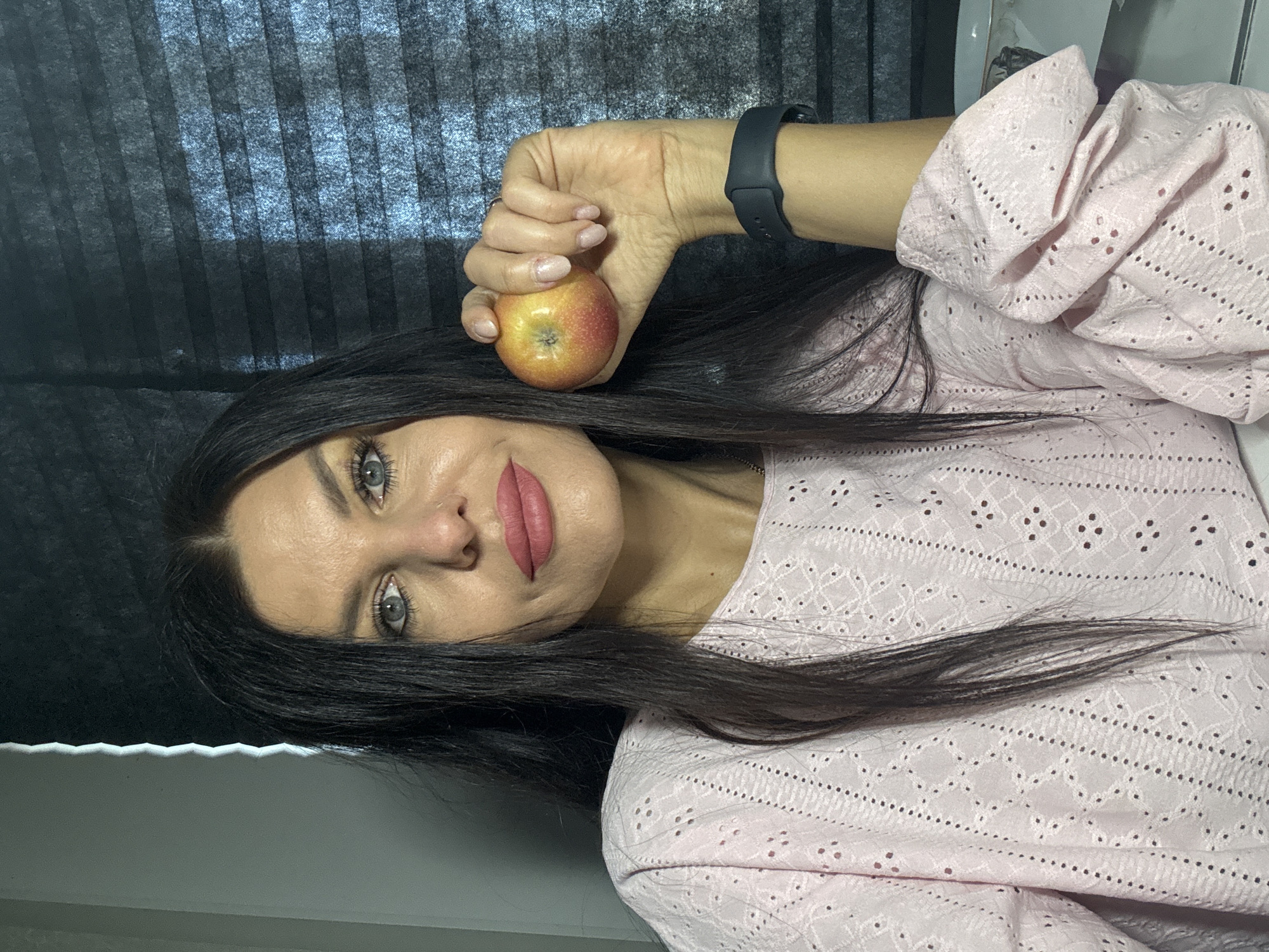 A portrait-style image, rotated 90 degrees counter-clockwise, features a woman with long, dark hair parted down the middle. She has prominent dark eyebrows and light blue or gray eyes, looking directly at the camera. Her lips are painted with pink lipstick. She is wearing a light pink, long-sleeved top or shirt with intricate eyelet or embroidered patterns. On her left wrist, she wears a dark-colored smartwatch or fitness tracker. The woman is holding a small, red and yellow apple in her left hand, which is positioned near her head.
The background consists of dark, vertically ribbed blinds or curtains, suggesting an indoor setting with natural light filtering through. A lighter, plain surface is visible in the lower right portion of the image. The woman has a neutral to slightly soft expression. The image's location is Chaikovsky, Russia. There is no visible text.