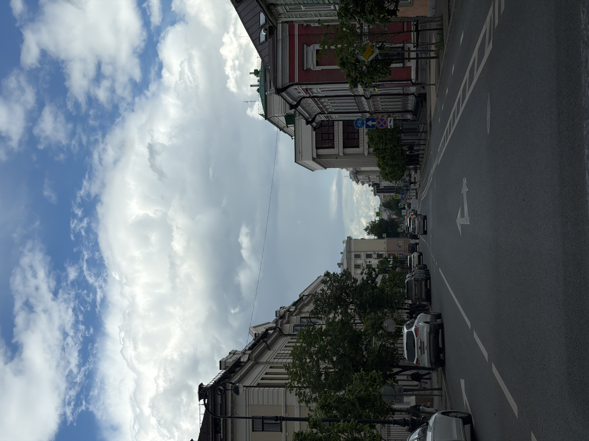 The image shows a street in Kazan, Russia, lined with buildings of varying architectural styles. The sky above is filled with clouds, with patches of blue visible. The street has multiple lanes of traffic with cars. Trees are planted along the sidewalks, partially obscuring some of the buildings. There are traffic signs visible, and street lighting is present. No people can be clearly identified.