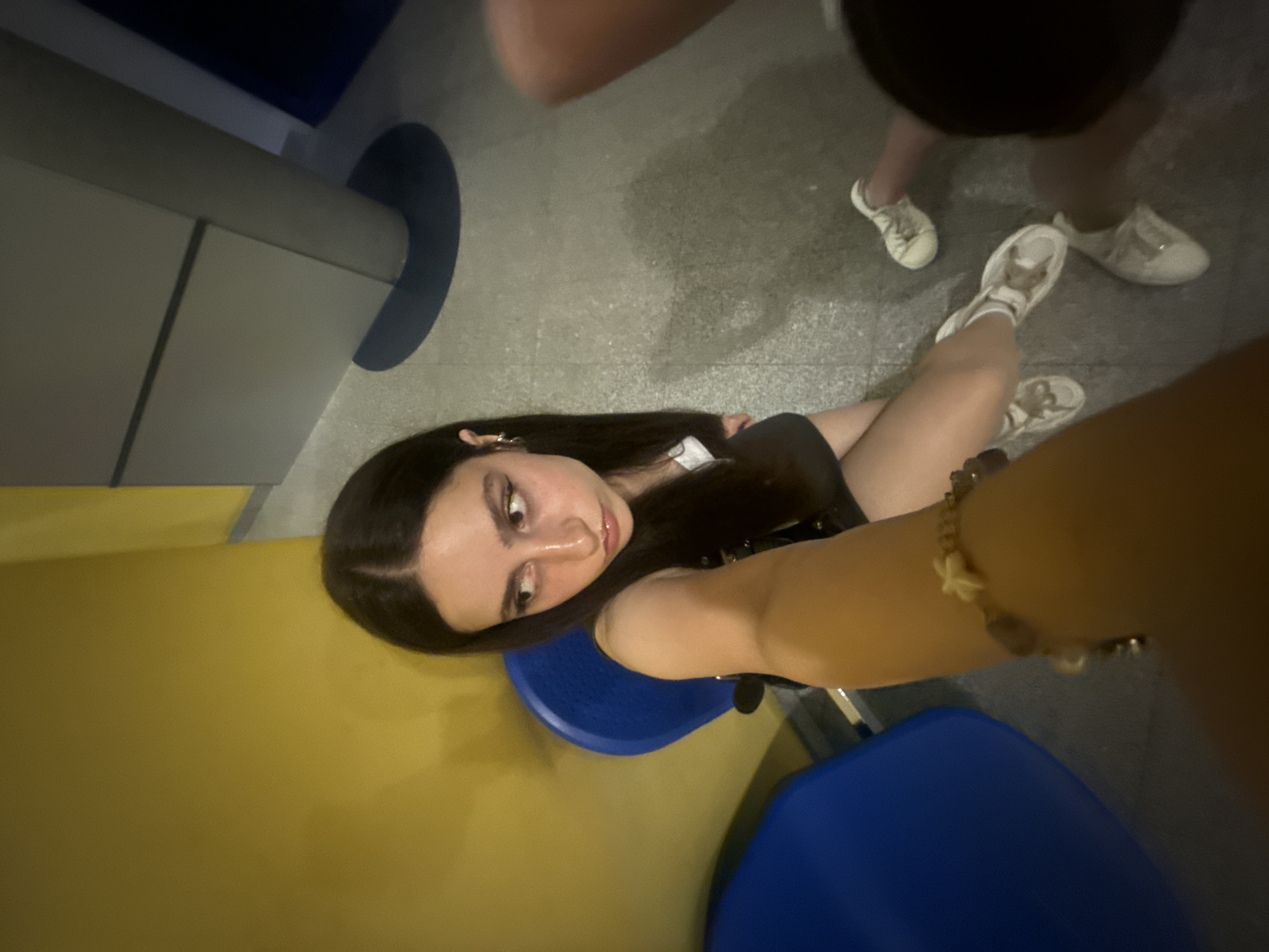A low-angle, wide shot shows a young woman with long, dark hair and notable makeup taking a selfie in what appears to be a public area. She is seated on a bright blue chair next to a yellow wall. The background consists of a tiled floor and architectural elements that suggest an indoor or covered public space. A second person's legs, wearing white sneakers, are visible in the lower right portion of the frame. The woman is wearing a dark top and a bracelet with several charms. Her facial expression is looking away from the camera. The lighting is somewhat dim, with shadows present on the floor. The image does not contain any discernible elements that would indicate the location is the city of Opole, Poland.