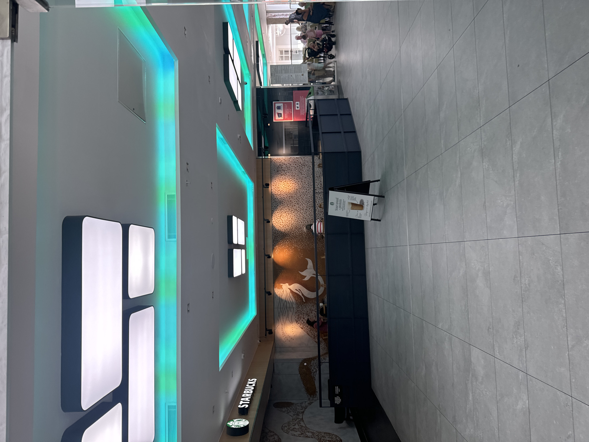 This image shows the interior of a Starbucks coffee shop, likely located in the city of Waterford, Ireland, as indicated by the context. The shop features modern lighting with illuminated ceiling elements in green and white. A prominent Starbucks logo with the siren illustration is visible on the wall behind the counter. The counter area is dark blue, and there are a few customers seated or ordering. In the foreground, a black A-frame sign displays an advertisement for a protein drink. The flooring is tiled in a light gray color. Through a glass partition to the right, a seating area with patrons, including families with children and strollers, is visible, suggesting a public or commercial space.