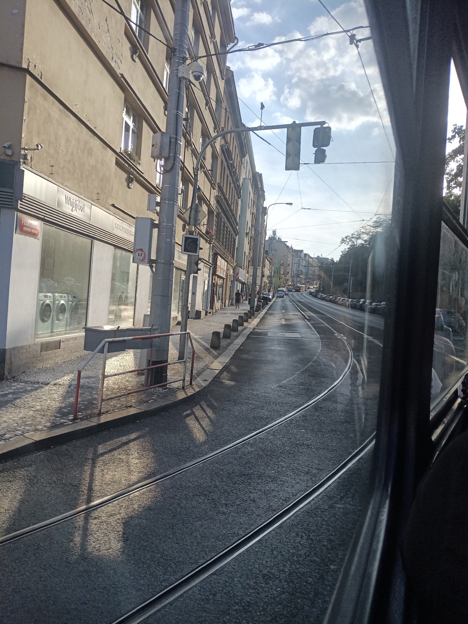 The image depicts a street scene in Prague, Czechia, viewed from the perspective of a moving tram. The street features tram tracks running along its length, and is lined with buildings on the left. The buildings are multi-story structures with a light-colored facade. One of the buildings has a storefront with large windows displaying washing machines, and signage for "Whirlpool". A traffic light hangs over the street. The street appears wet, possibly from recent rain, with reflections visible on the asphalt. parked cars line the right side of the street. The sky is partly cloudy. The overall impression is that of an urban environment on a bright day.