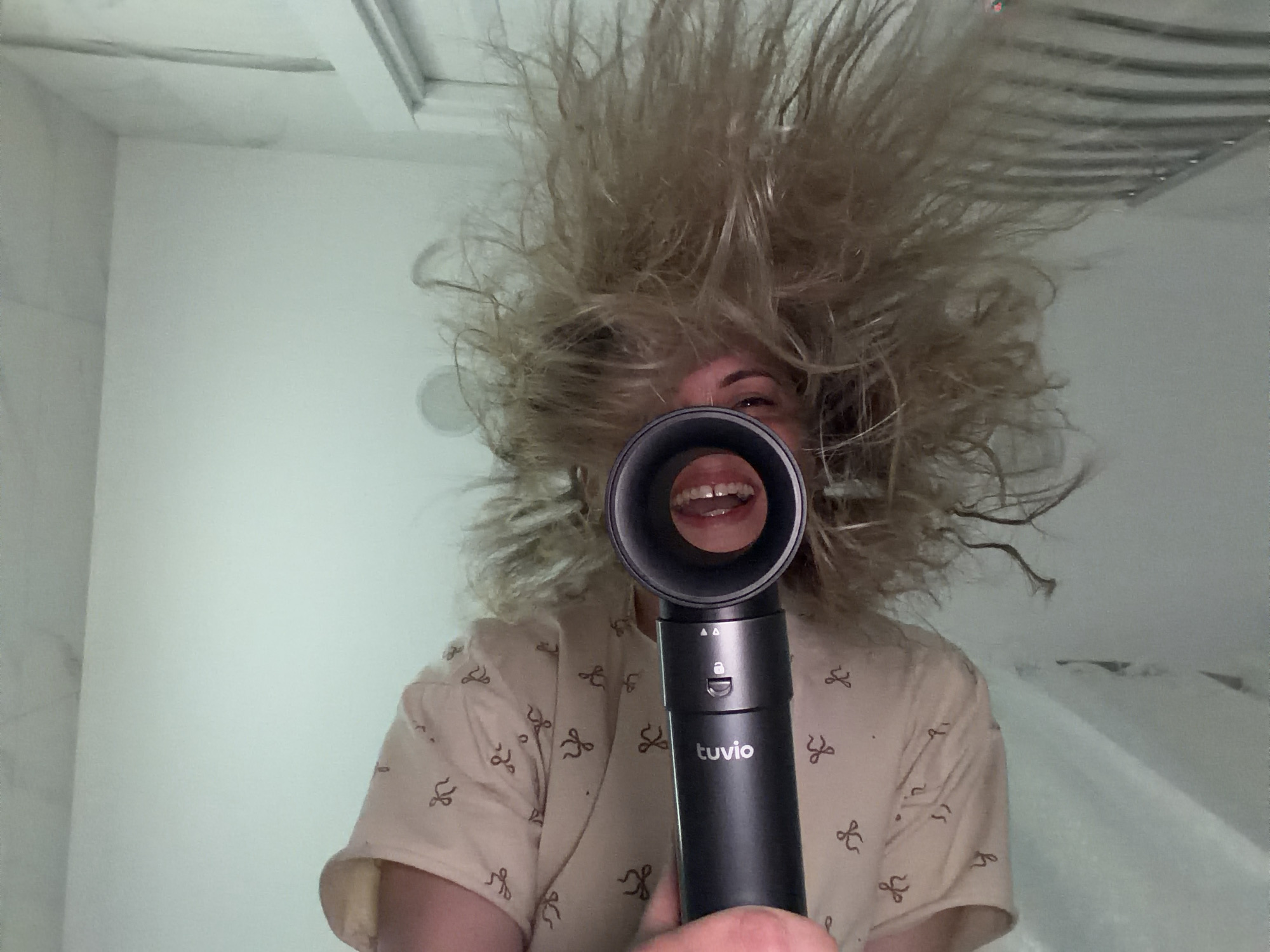 A person with dramatically wild, frizzy blonde hair is the central subject of this indoor, head-on shot, taken in Votkinsk, Russia. The person holds a black, sleek hair styling device, branded "tuvio," directly in front of the camera, creating a humorous visual where their smiling mouth and teeth are visible through the device's circular opening. Their hair is extremely voluminous and stands on end, as if affected by powerful airflow or static electricity, suggesting they are either actively using the device, have just finished using it, or are playfully posing to exaggerate the effect.
The person is wearing a light-colored, possibly beige or light pink, short-sleeved top adorned with numerous small, dark brown bow-tie patterns. The "tuvio" device features the brand name prominently in white text, along with small white icons including what appears to be a lock symbol and two arrow indicators on its handle.
The background consists of plain white walls and a white ceiling, which shows some subtle linear details, possibly from ceiling panels or a ventilation grate. The lighting is bright and even, consistent with an indoor setting like a bathroom or a well-lit room. The image captures a lighthearted, playful moment, emphasized by the person's smile and dramatically styled hair.