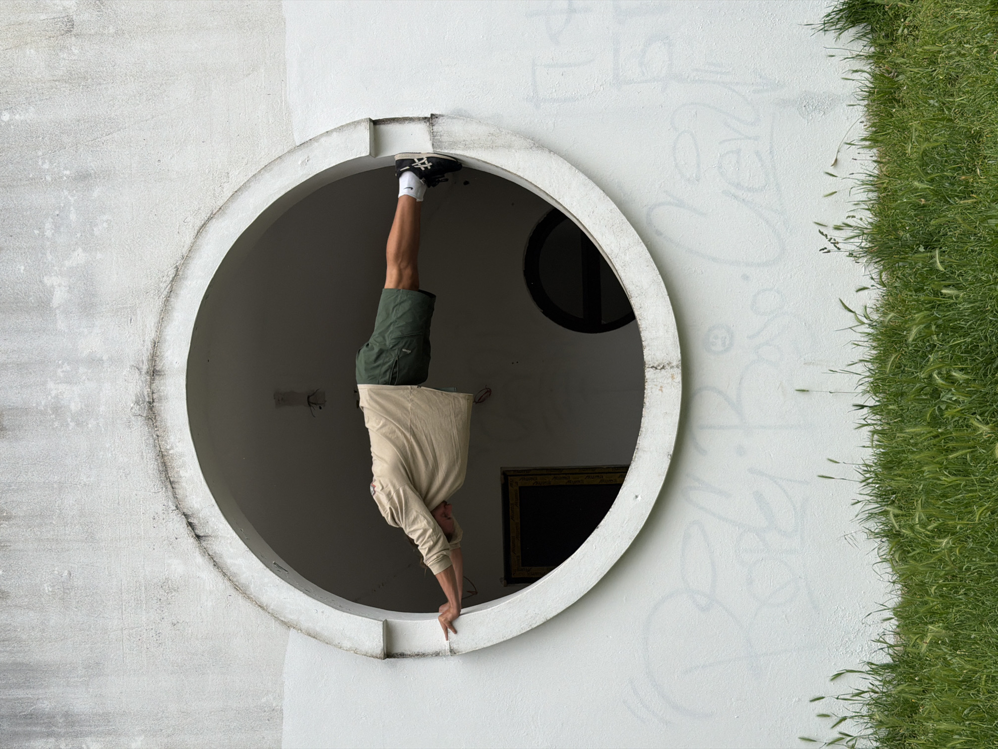 A person is upside down, standing on his hands in a circular window opening of a white building, with his feet in the top of the circular window. The person is wearing a beige long-sleeved shirt, green shorts, white socks, and black shoes. The building is located in Petrovac centar, Montenegro. Grass is growing up the side of the building on the right side of the image. There are two more dark, circular openings inside the building, and one framed painting. There is graffiti on the white wall.