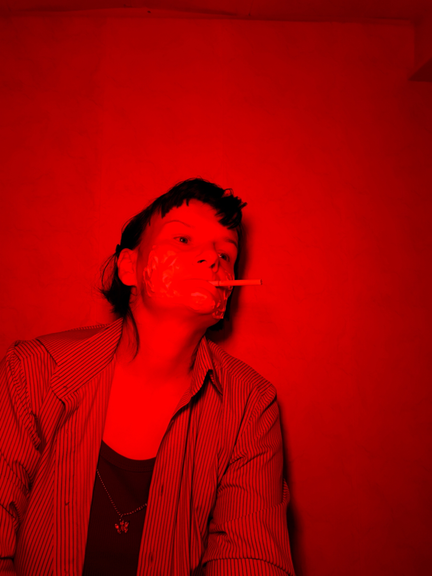 A person stands against a solid red wall, lit by red light. The person is wearing a striped shirt over a dark tank top and a necklace with a small pendant. They have dark hair and appear to have a substance smeared across their face. A cigarette is held in their mouth. The location in the city of Фоки, Russia, is not evident in the image.
