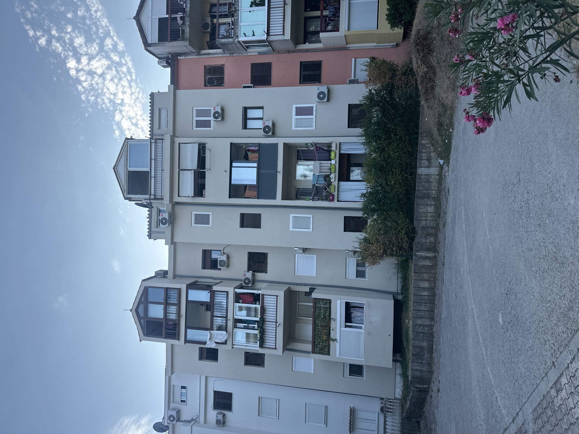 The image depicts a residential building in Budva, Montenegro. The building is light beige with several balconies and windows. Some balconies have clothes hanging out to dry, and air conditioning units are visible on the exterior walls. There are also some trees and bushes in front of the building, and a paved area in the foreground. The sky is cloudy and a light blue. The overall scene is one of a typical residential neighborhood.