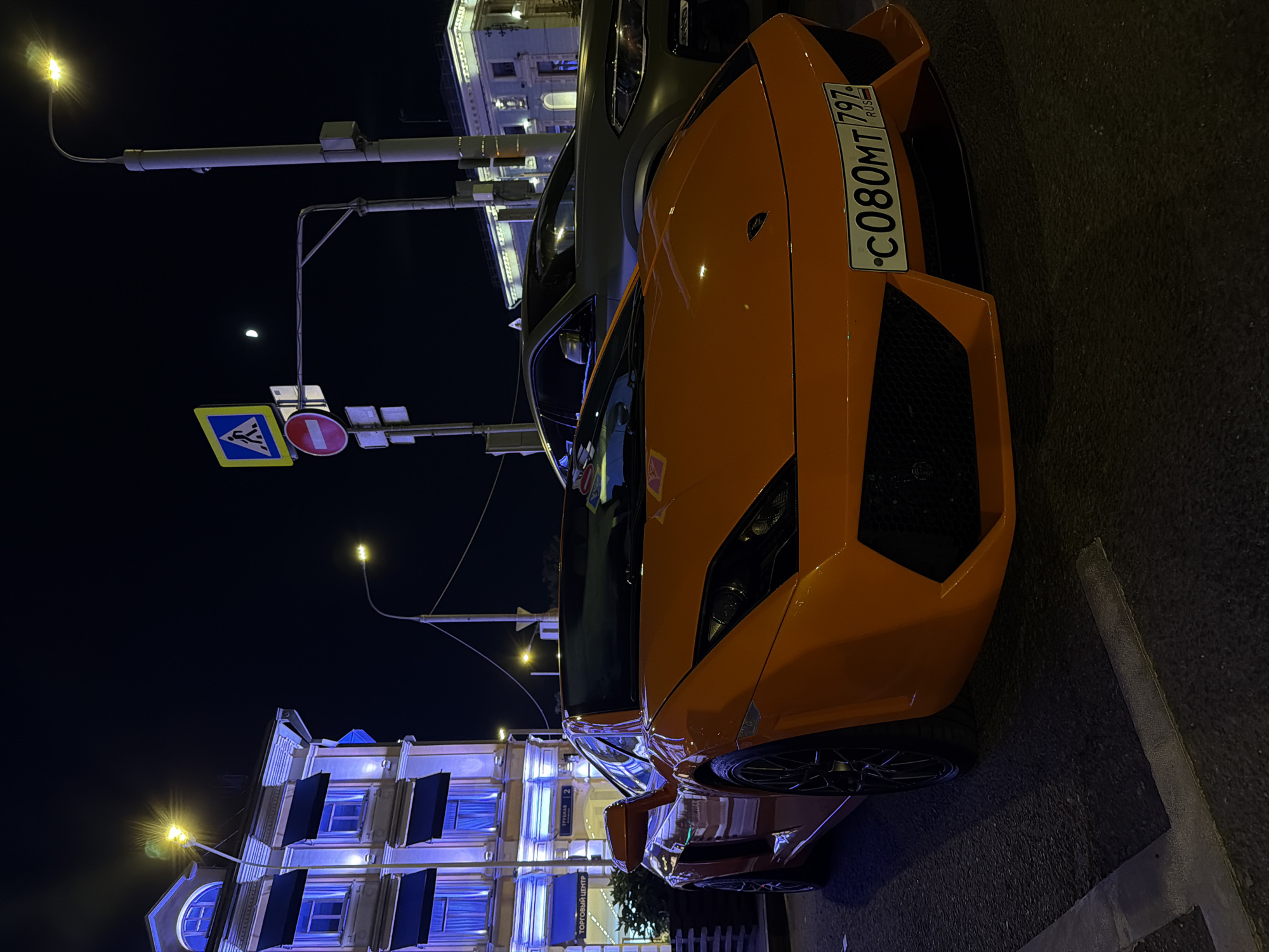 At night in Moscow, Russia, an orange Lamborghini with license plate "C080MT 797" is parked on a city street. The background includes illuminated buildings, streetlights, and a crosswalk sign.