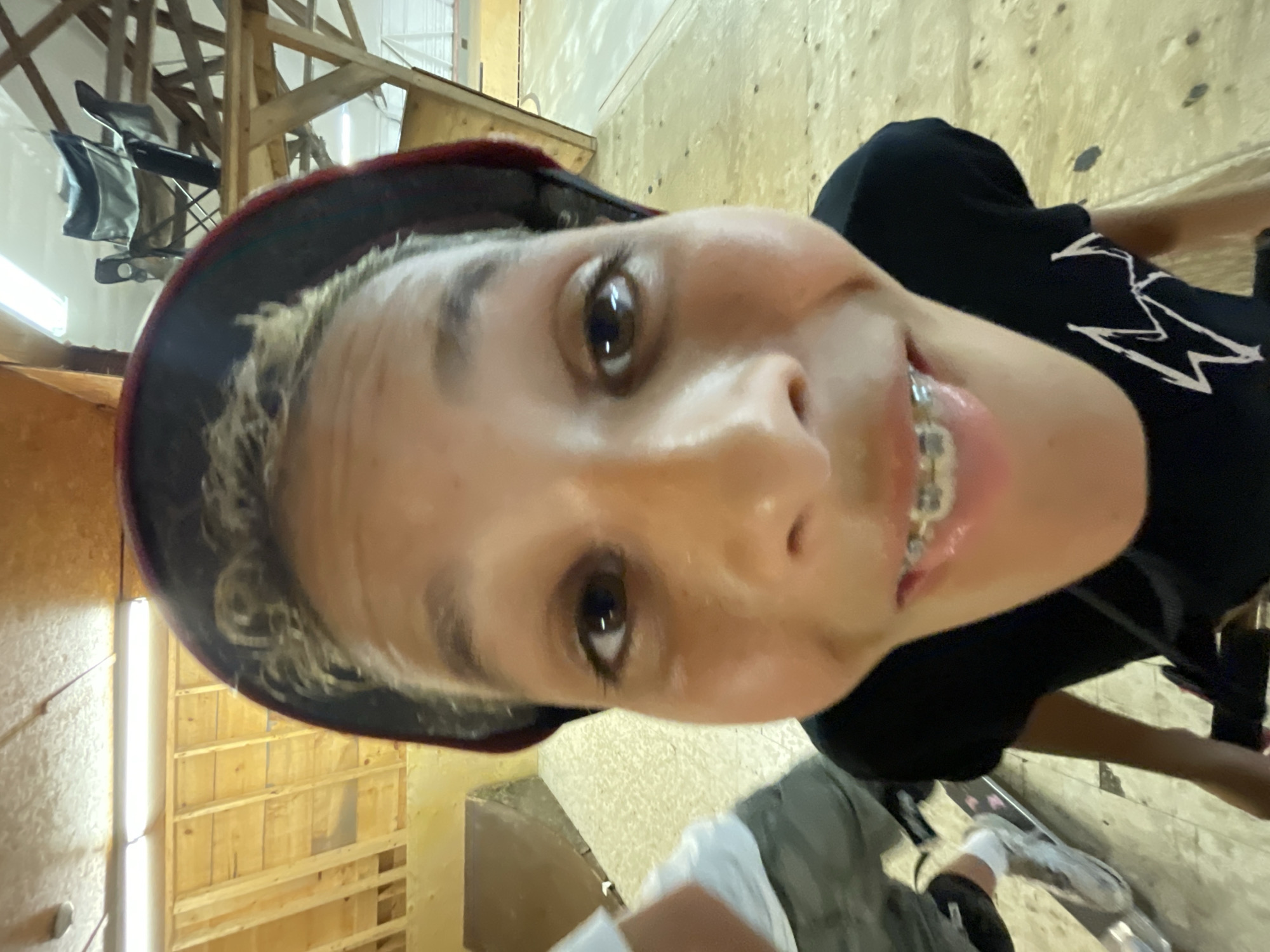 The image shows a close-up, fisheye lens shot of a young person wearing a helmet and a black t-shirt. The person has braces and is smiling. The background appears to be an indoor skate park with wooden ramps and a wooden floor. The lighting suggests it is daytime. The location is not specified in the image.